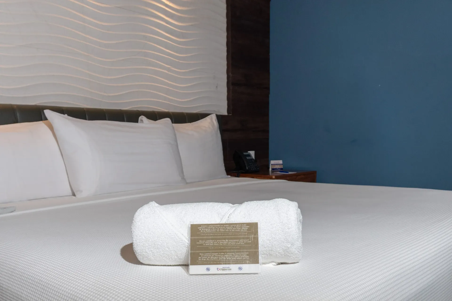 Una toalla blanca enrollada con una pequeña tarjeta reposa sobre una cama con almohadas blancas, frente a una pared blanca texturizada y una pared azul.