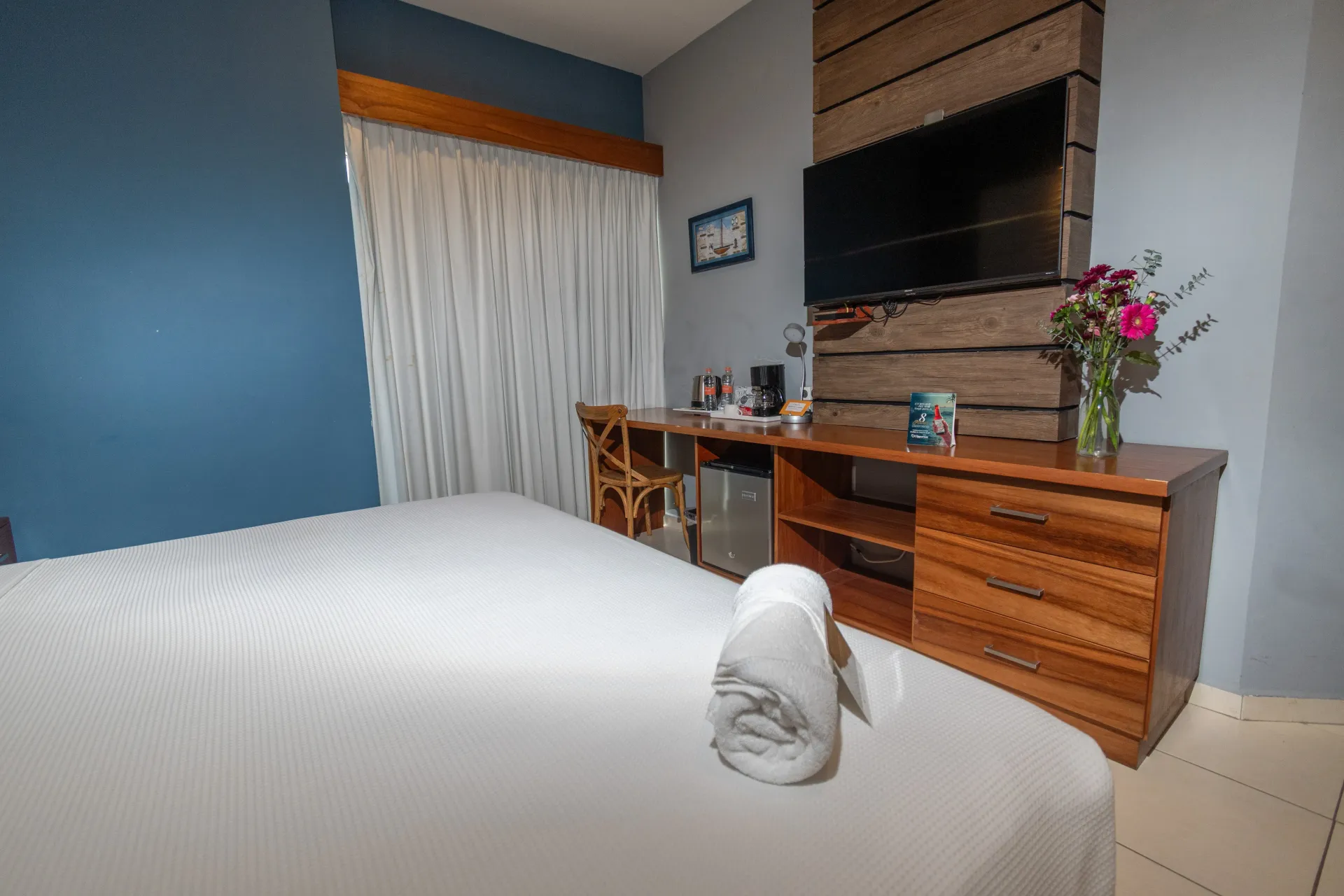 Un dormitorio moderno con una cama con sábanas blancas, una cómoda de madera con un televisor montado y una pared decorativa de color azul.