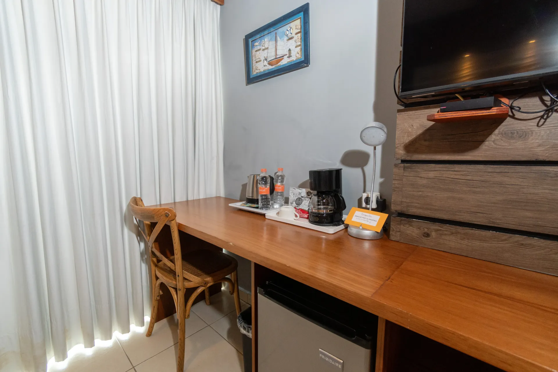 Escritorio de madera con cafetera y lámpara  junto a una silla y cortinas blancas, debajo de una tv, cuadros en la pared