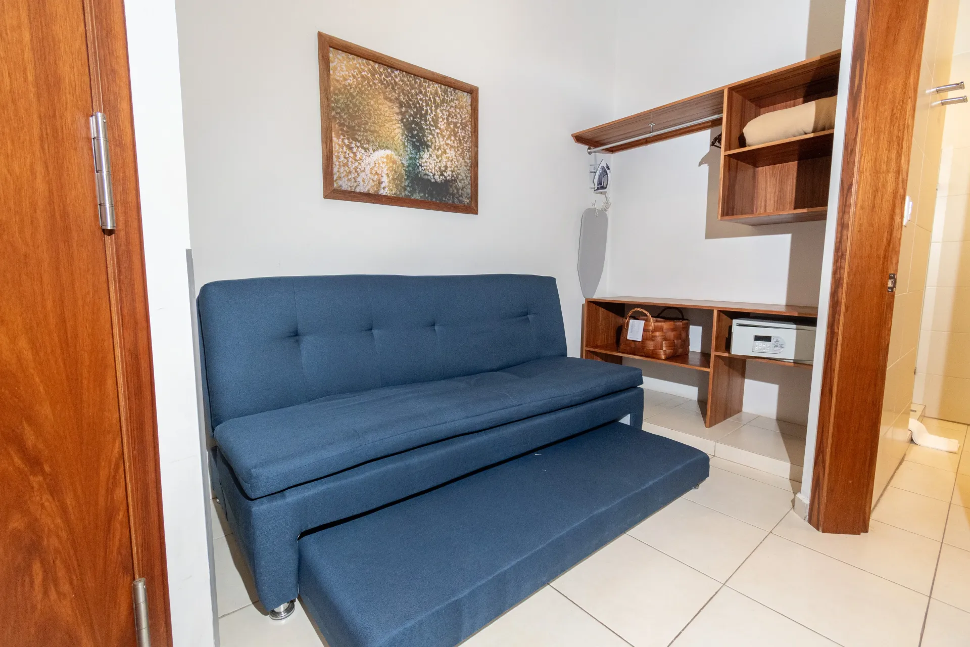 Sofá cama azul en una habitación con suelo de baldosas de color claro y estantes de madera montados en la pared.