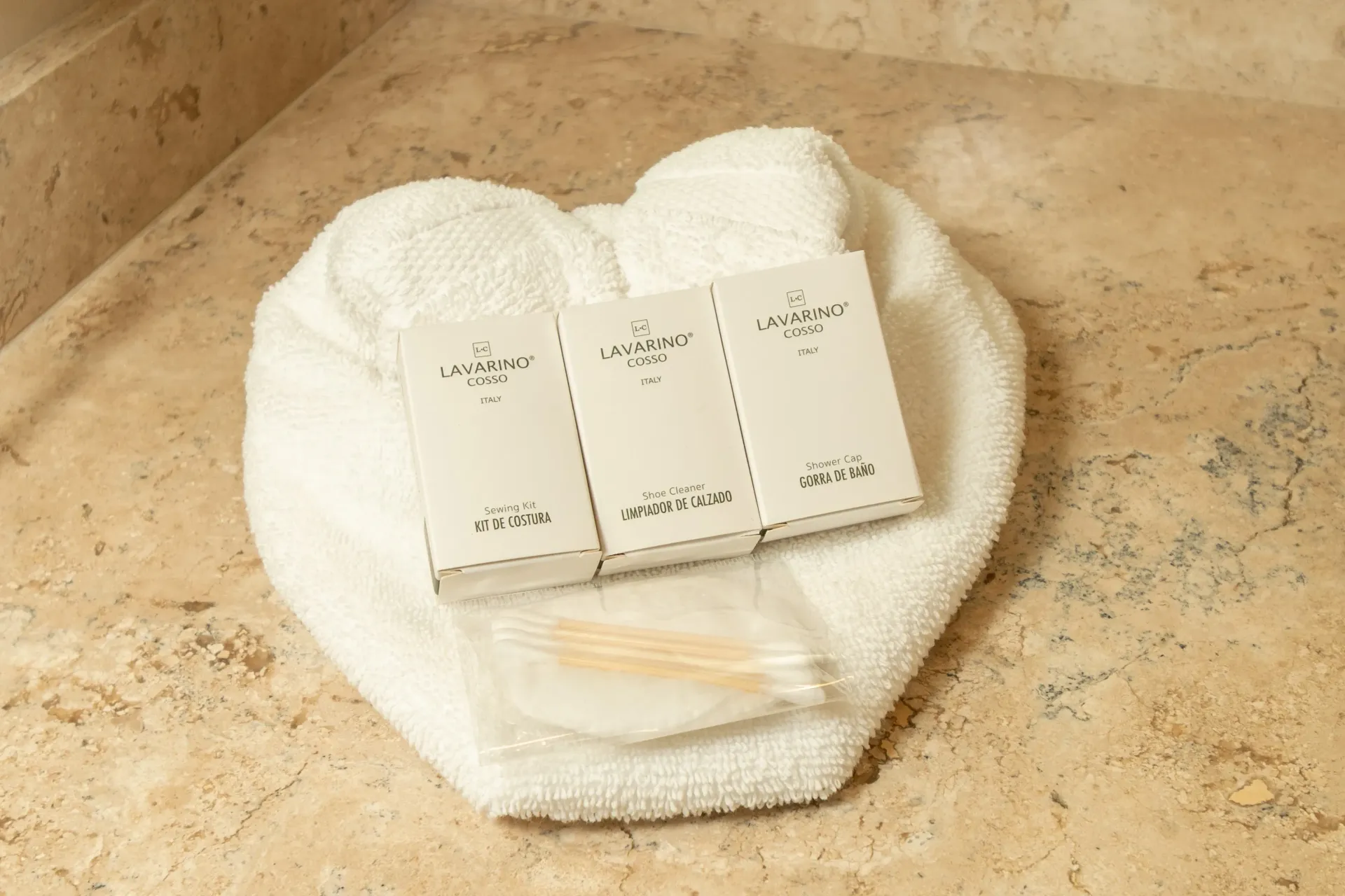 Three hotel toiletries in white boxes rest on a folded white towel with a small packet of cotton swabs on a stone surface.