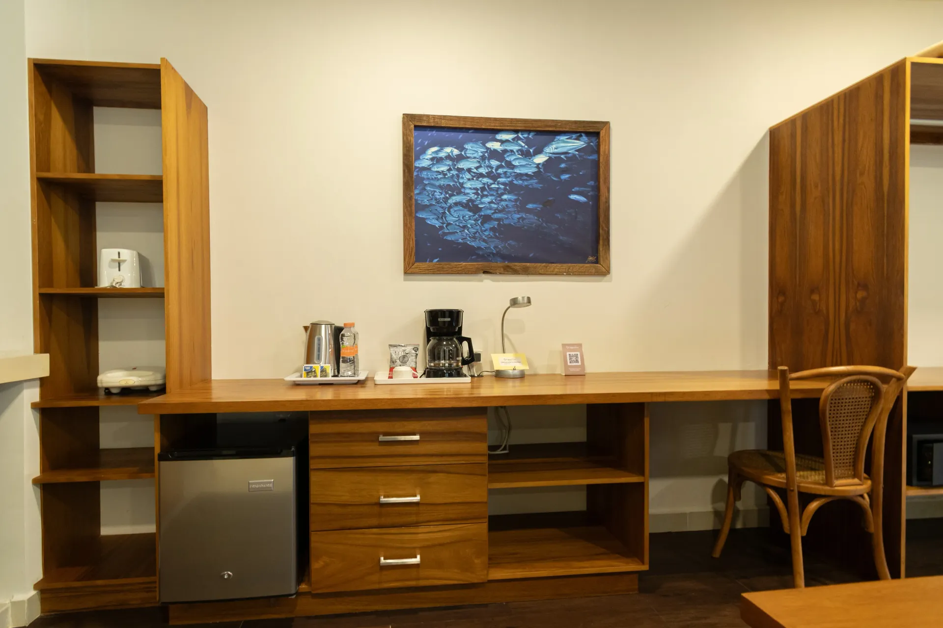 A wood desk unit with shelves, a mini-fridge, coffee maker, and a wooden chair against a plain wall.