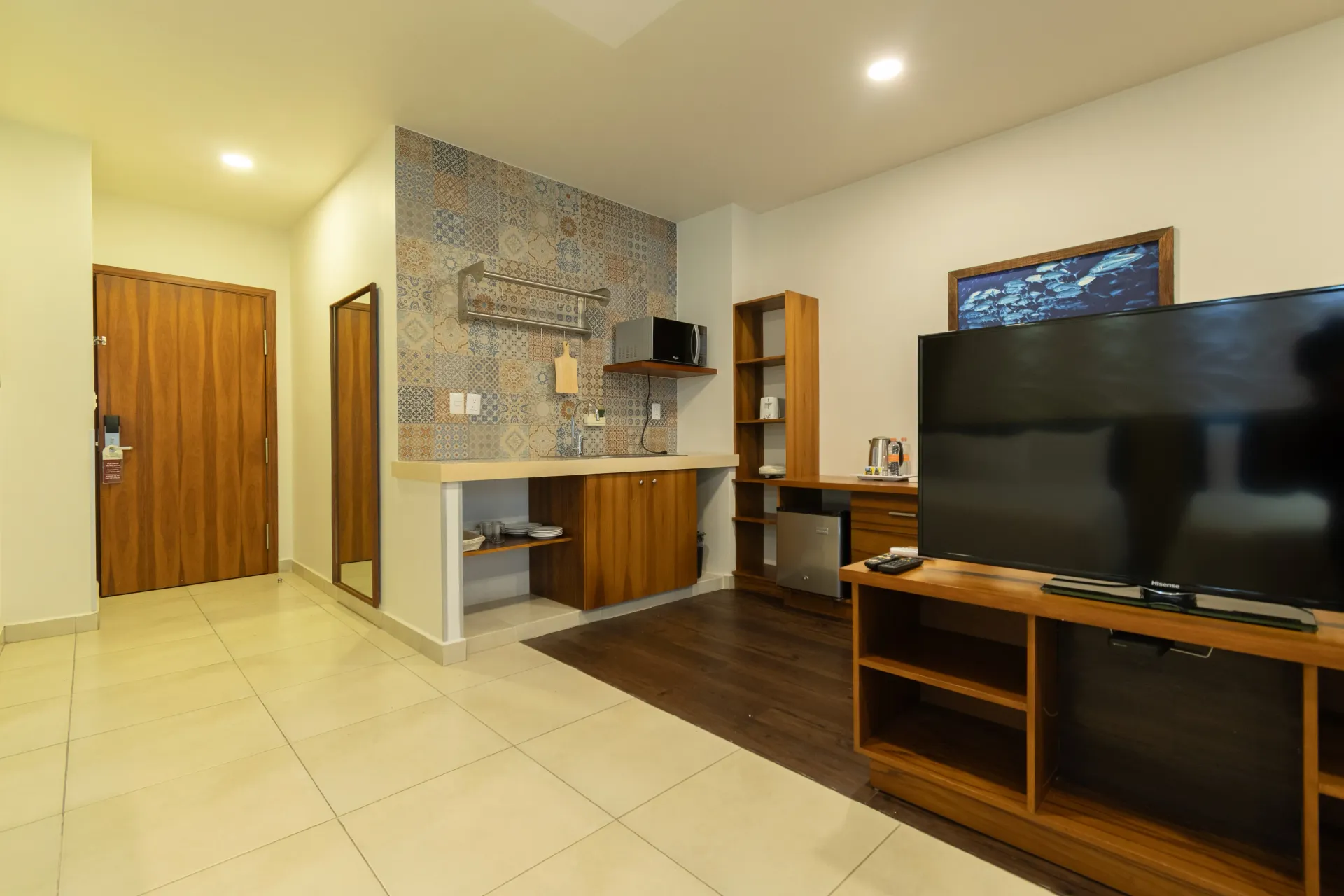 A hotel room kitchenette with patterned tile backsplash, wooden cabinets, a TV console, and an entryway door.