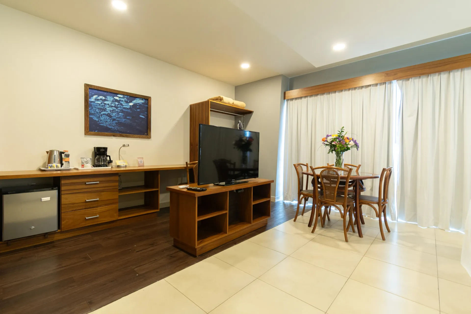 A hotel room with a dining table, a television on a wooden console, and a kitchenette area with a mini-fridge.