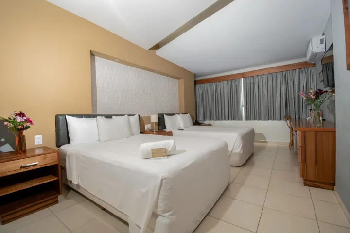 Habitación de hotel con dos camas blancas, mesita de noche de madera con flores y ventanales que van del suelo al techo 