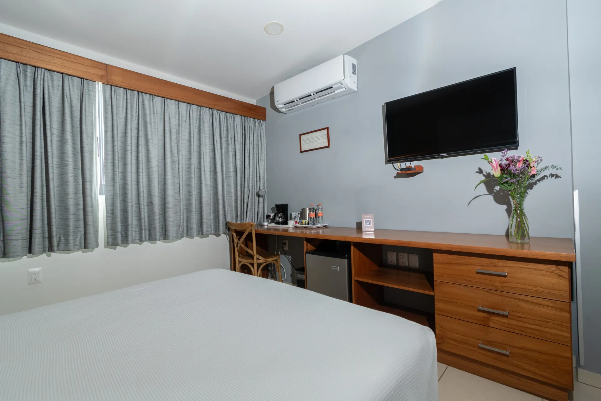 A hotel room with a bed, gray curtains, a wall-mounted TV, and a wooden desk with a small refrigerator and flower vase.