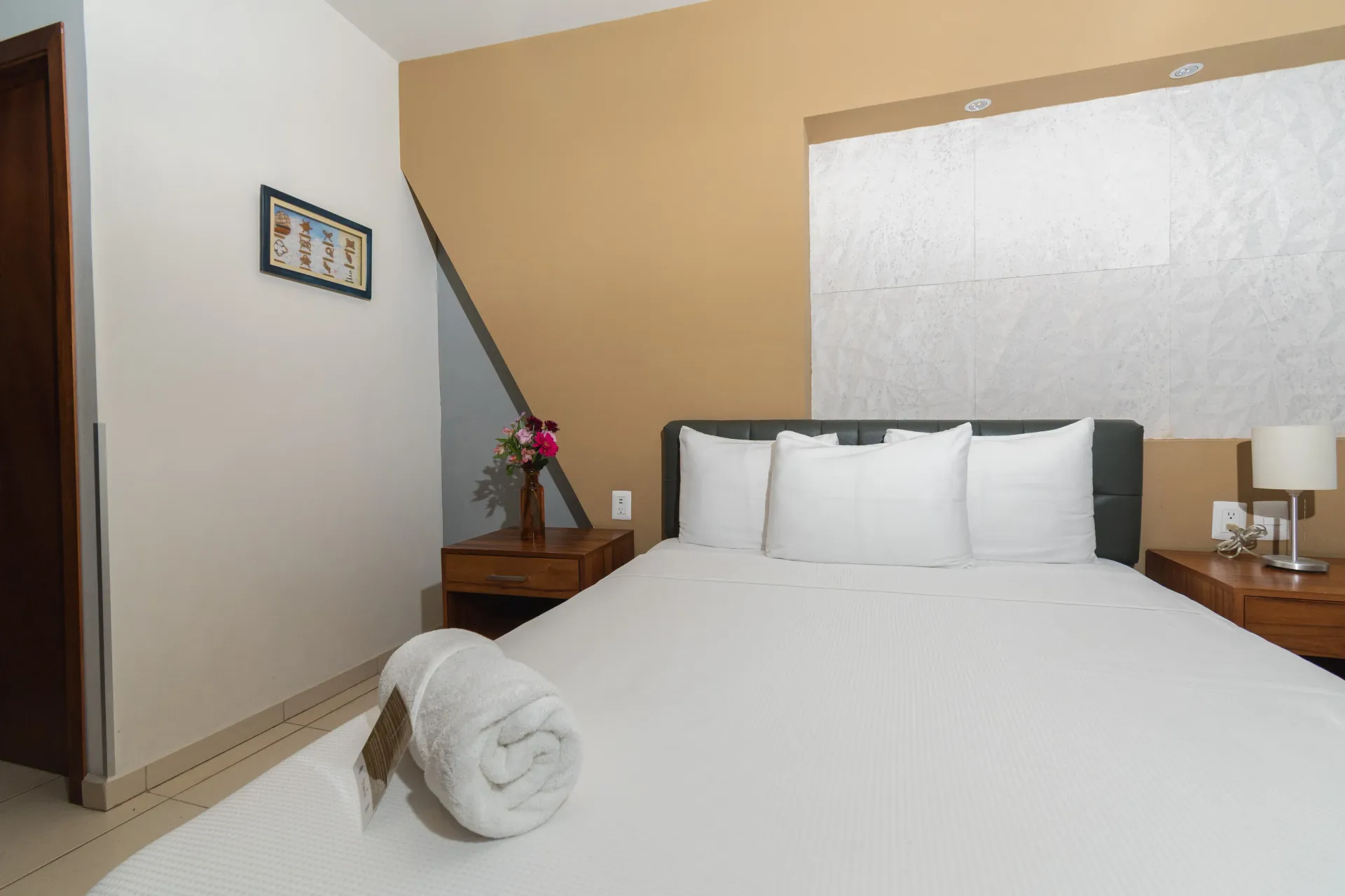 A neatly made bed with white linens and a rolled towel in a bedroom featuring beige walls, gray accents, and a nightstand.