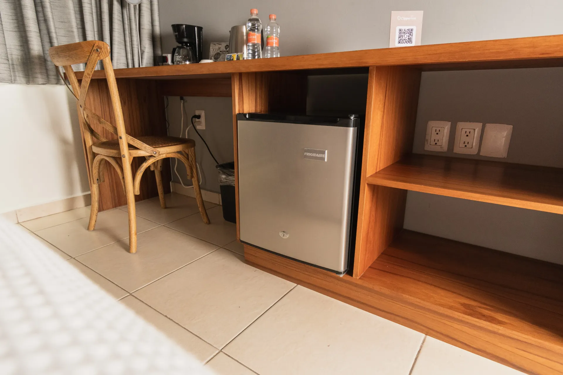 A wooden desk with a chair, small silver refrigerator, coffee maker, and water bottles against a light wall.