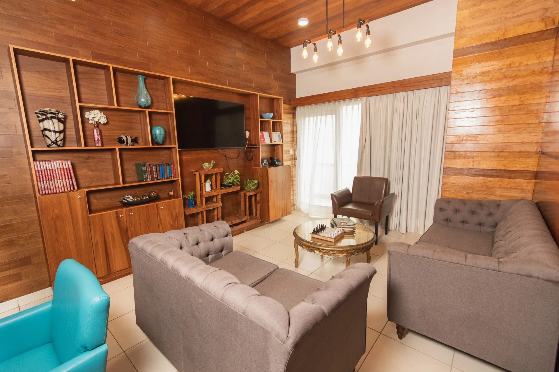 Sala de estar con paredes y estanterías de madera, TV, dos sofás grises, un sillón marrón y una silla color turquesa.