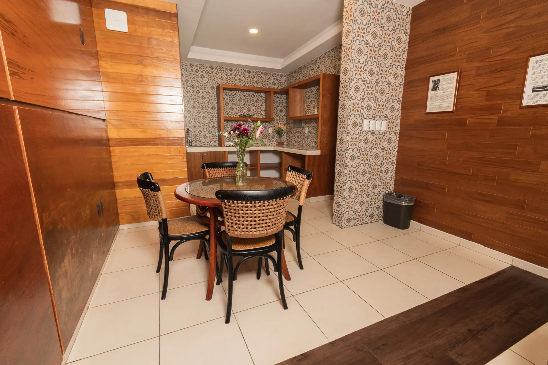 Comedor con una mesa redonda de cristal y cuatro sillas de mimbre, en una habitación con paredes revestidas de madera 