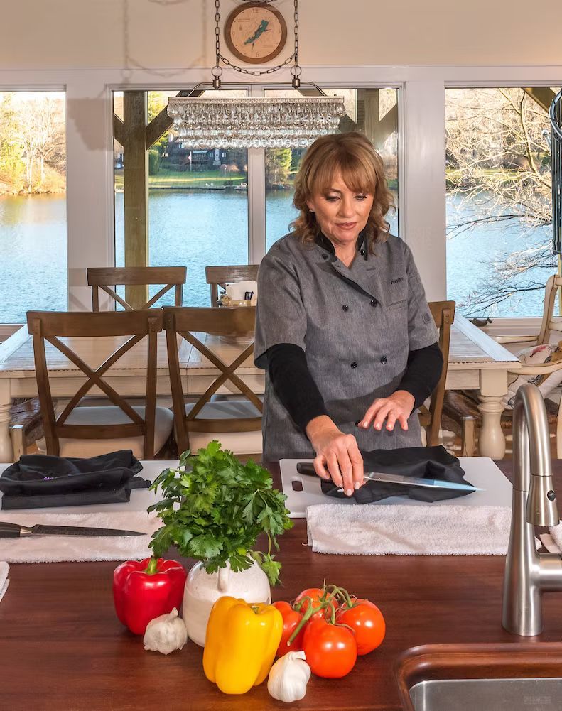 Woman arranging folded cloths on a table with peppers and herbs in a bright kitchen overlooking water