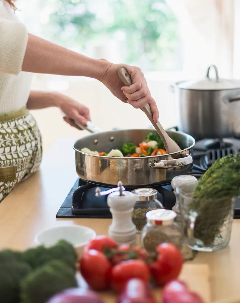 Person stirring vegetables in a pan on a stovetop with herbs and tomatoes in the foreground