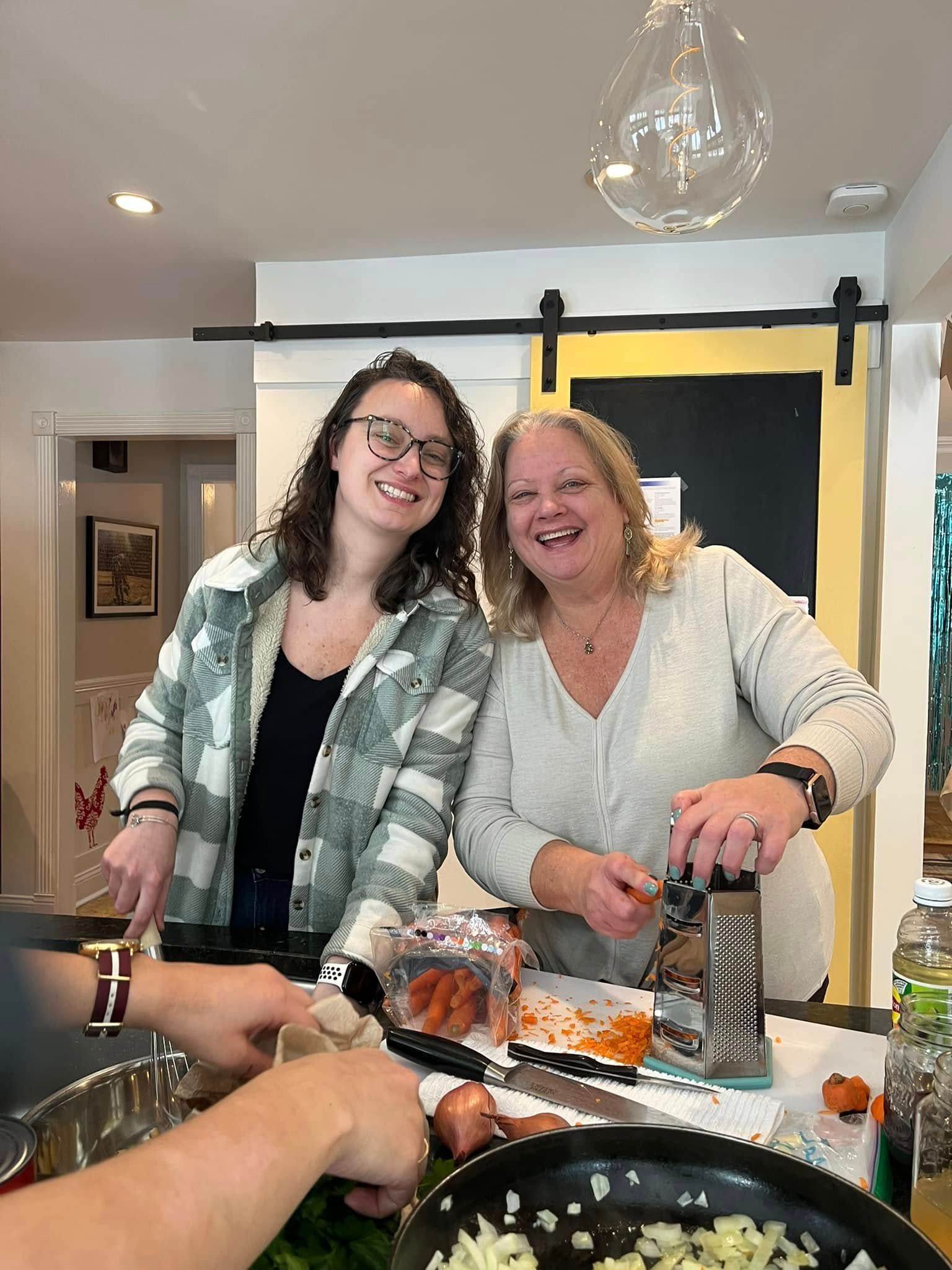 Two women smiling at a dining table, with wine bottles and dishes in a warmly lit room.
