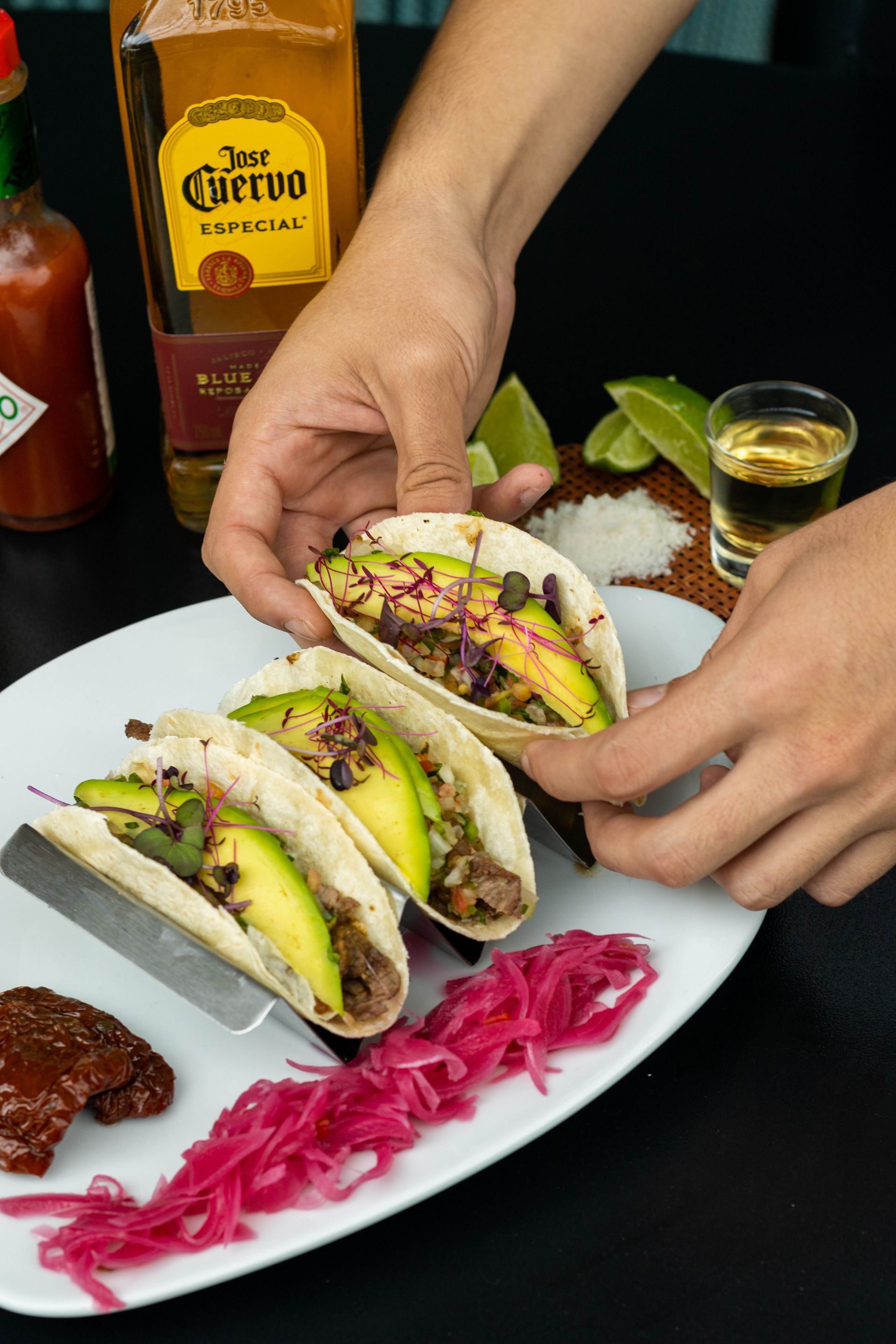 Hands arranging three avocado tacos on a white plate with lime and pickled onions