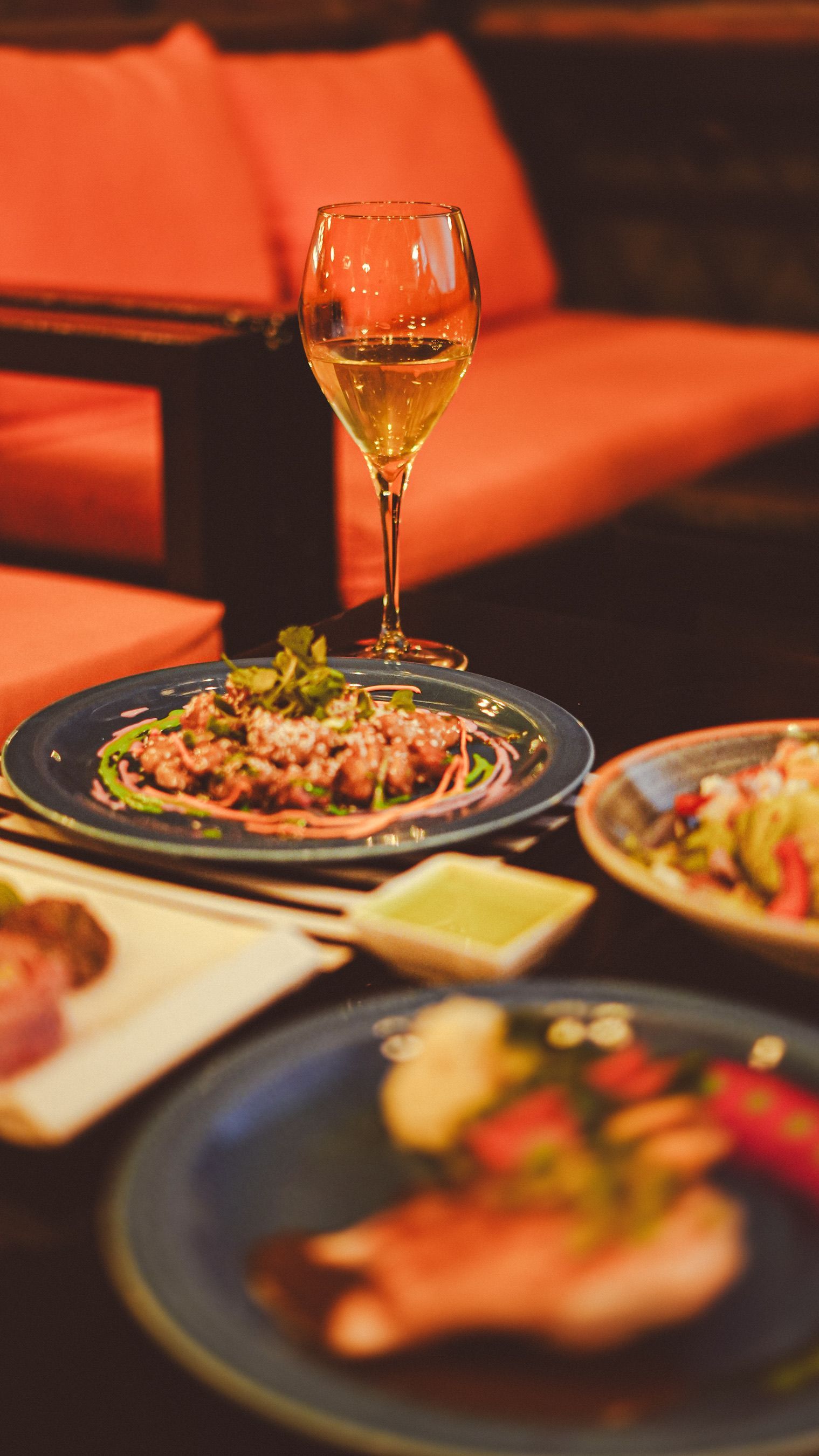 Restaurant table with a wine glass and plated food on a dark blue dish, in front of an orange sofa.