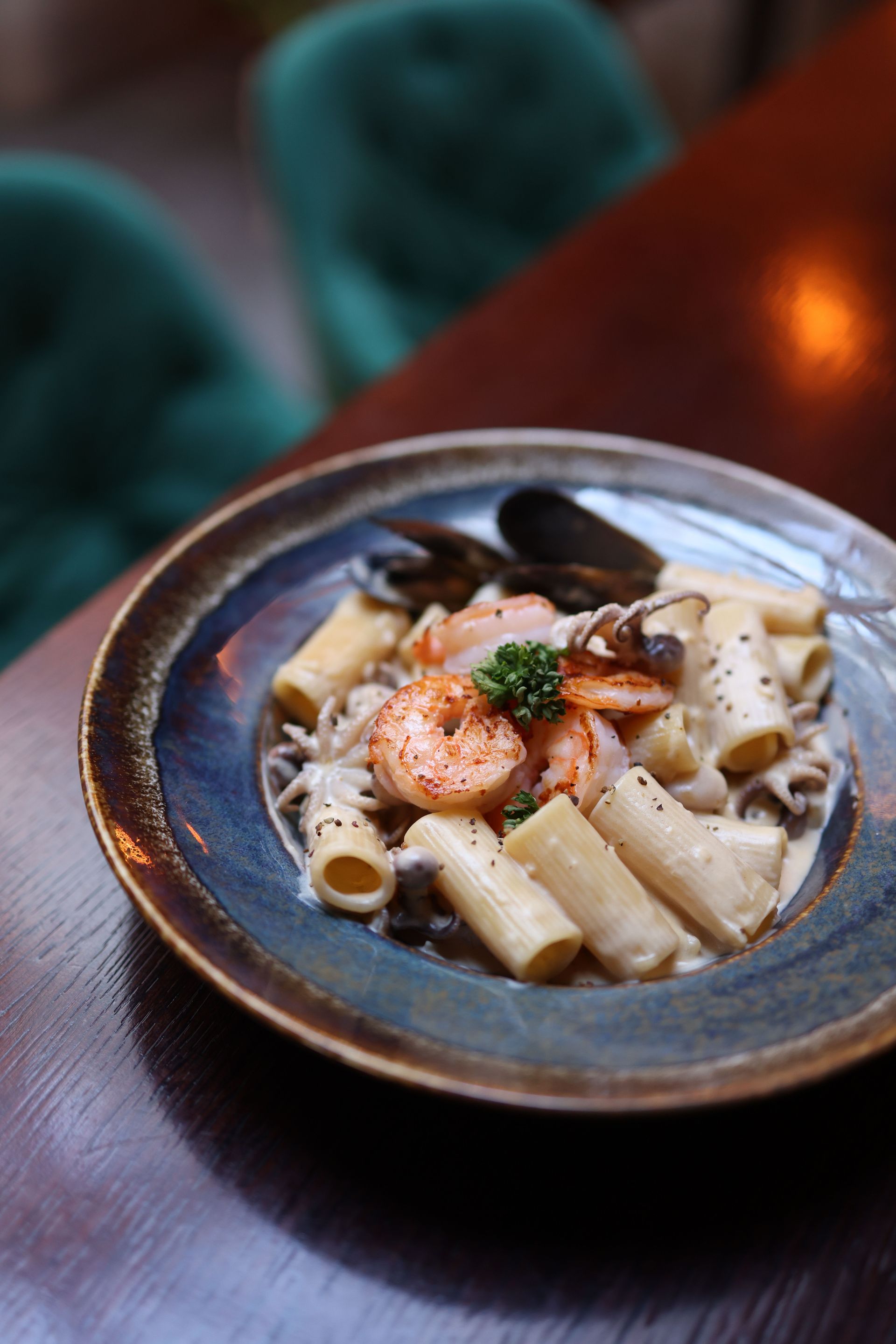 Pasta with shrimp in a dark bowl on a wooden table, with blurred green chairs in the background