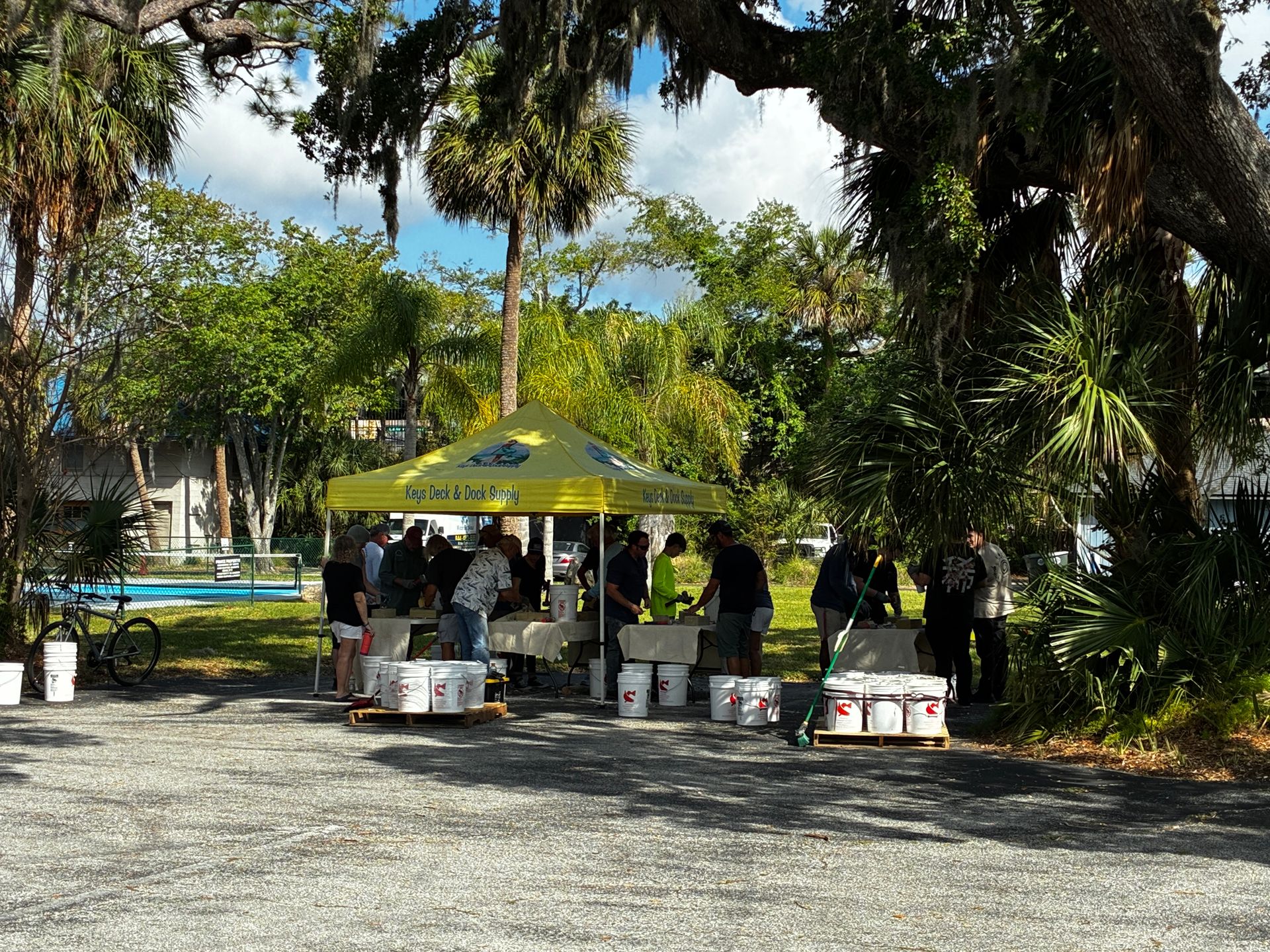 People gathered under a yellow canopy in a shady park with palm trees and picnic tables