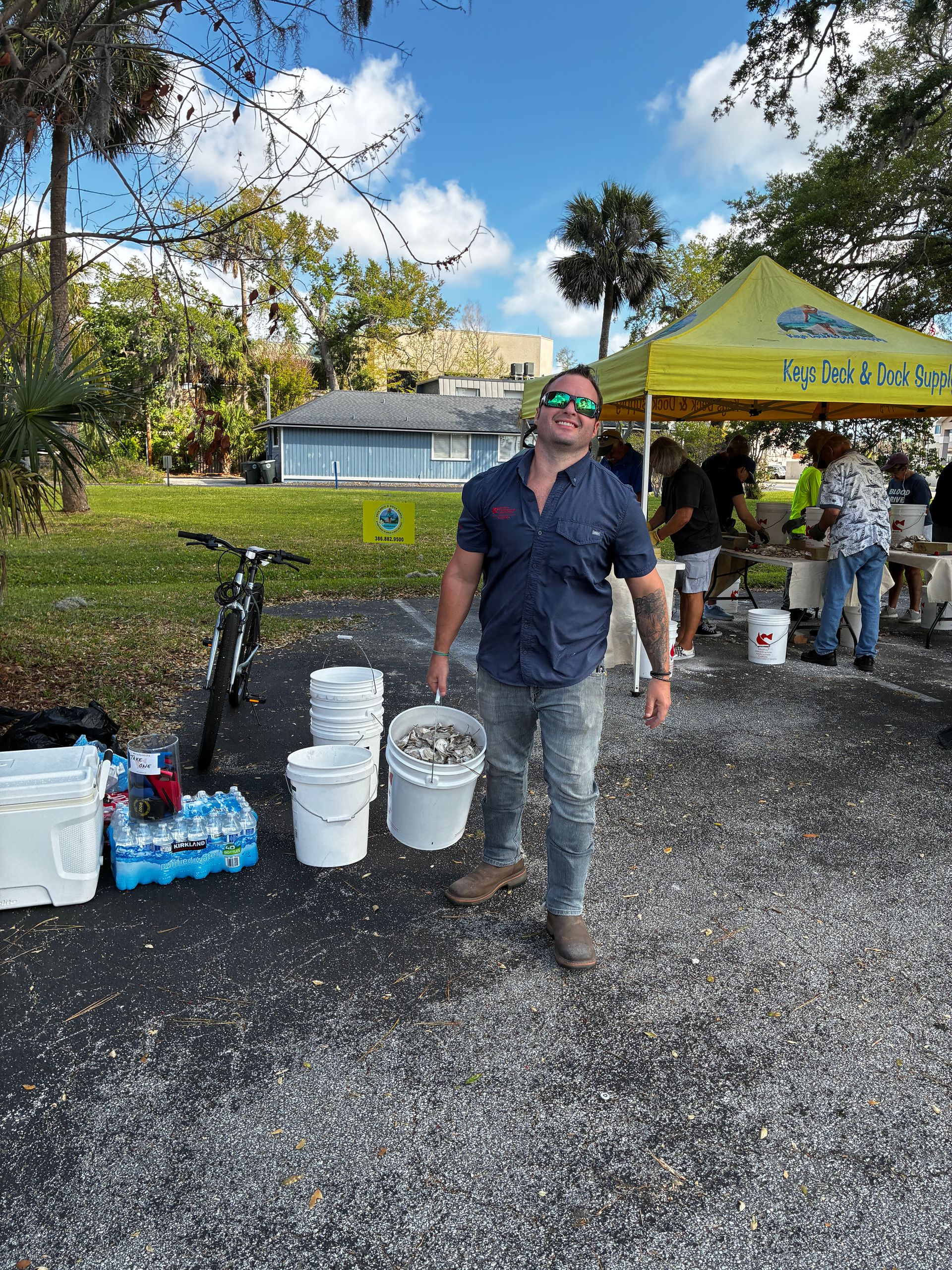 Person standing beside buckets and a bicycle at an outdoor drink station under a yellow tent.