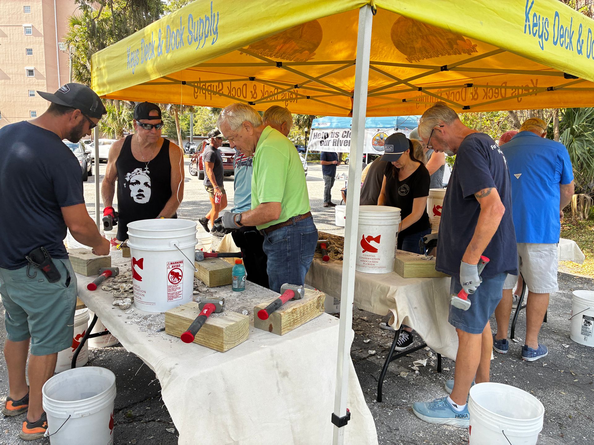 Volunteers serving food at an outdoor street fair under a yellow canopy with coolers and buckets