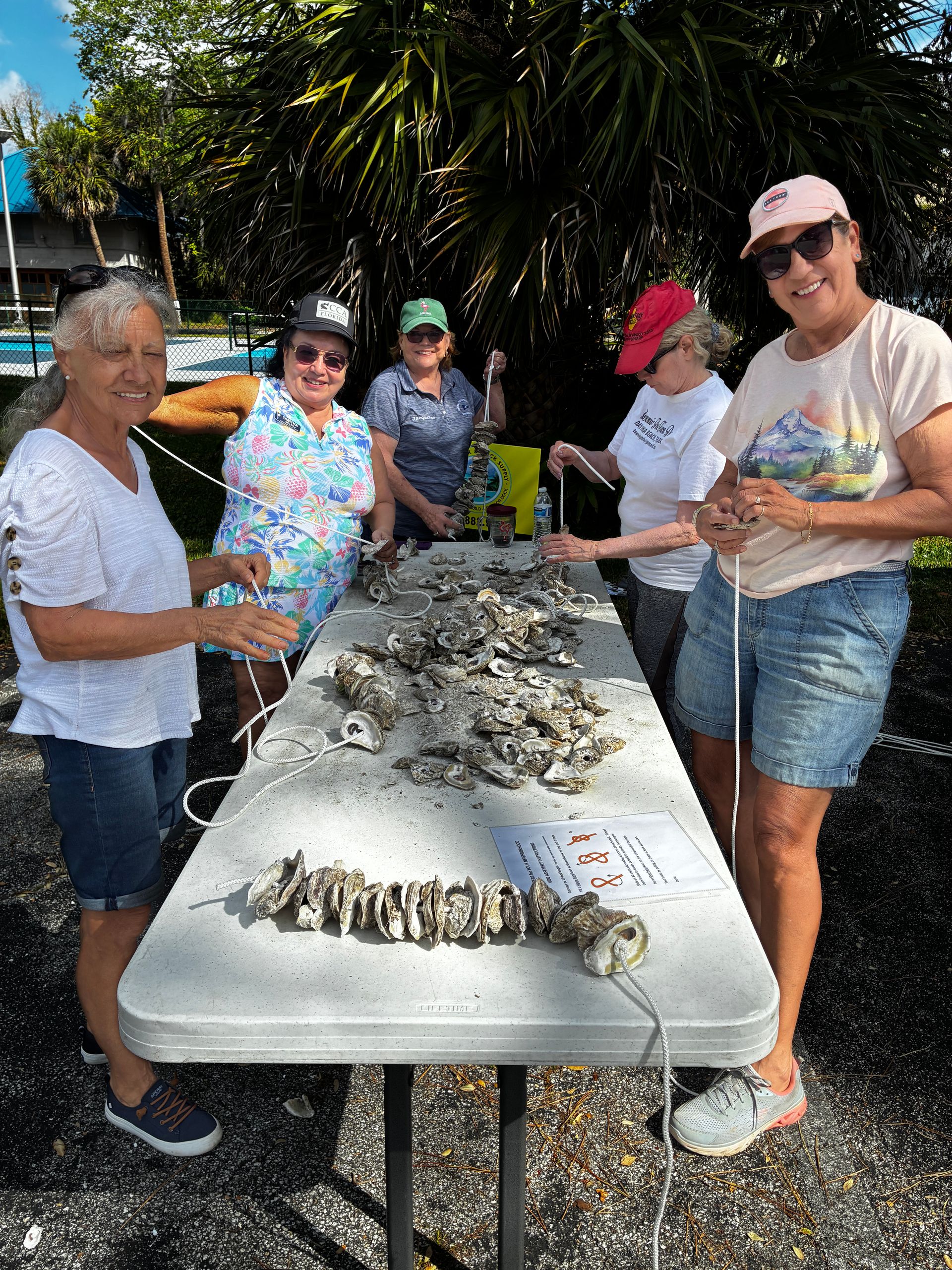 People standing around a table covered with piles of shells outdoors in bright sunlight