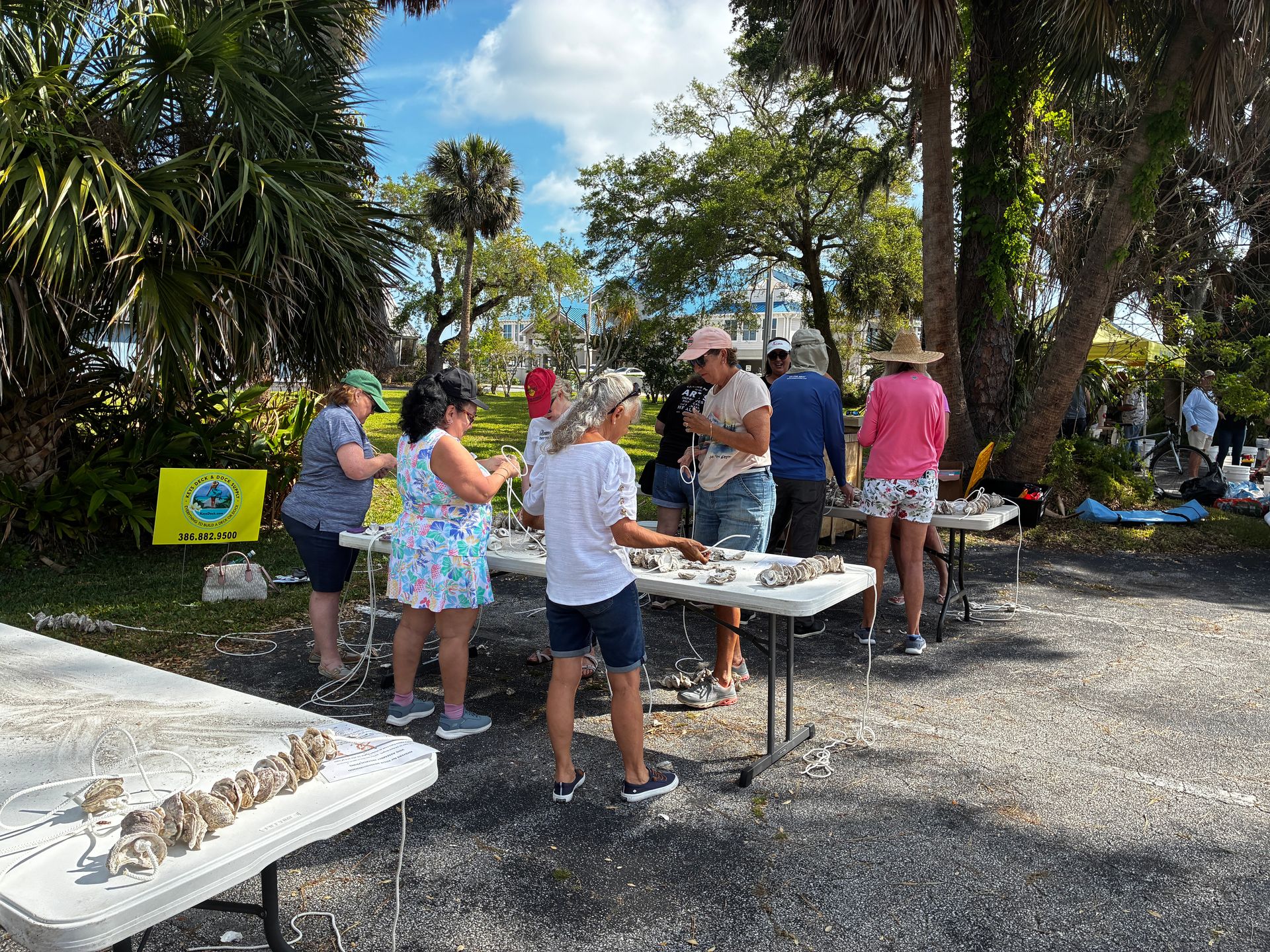 People gathered at outdoor picnic tables under trees, socializing at a sunny park event.