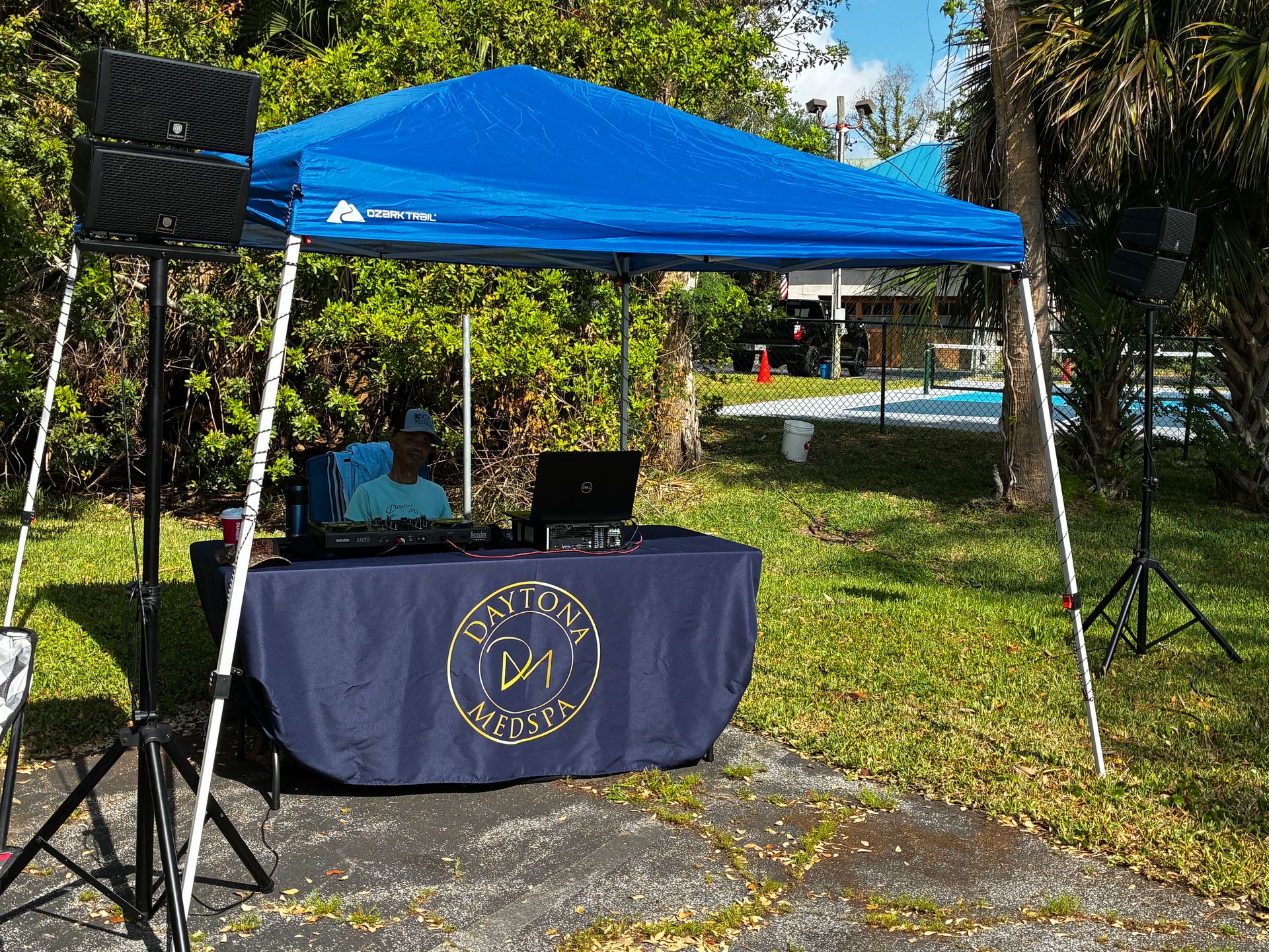 Blue canopy tent with table and speakers set up on a grassy outdoor lawn.