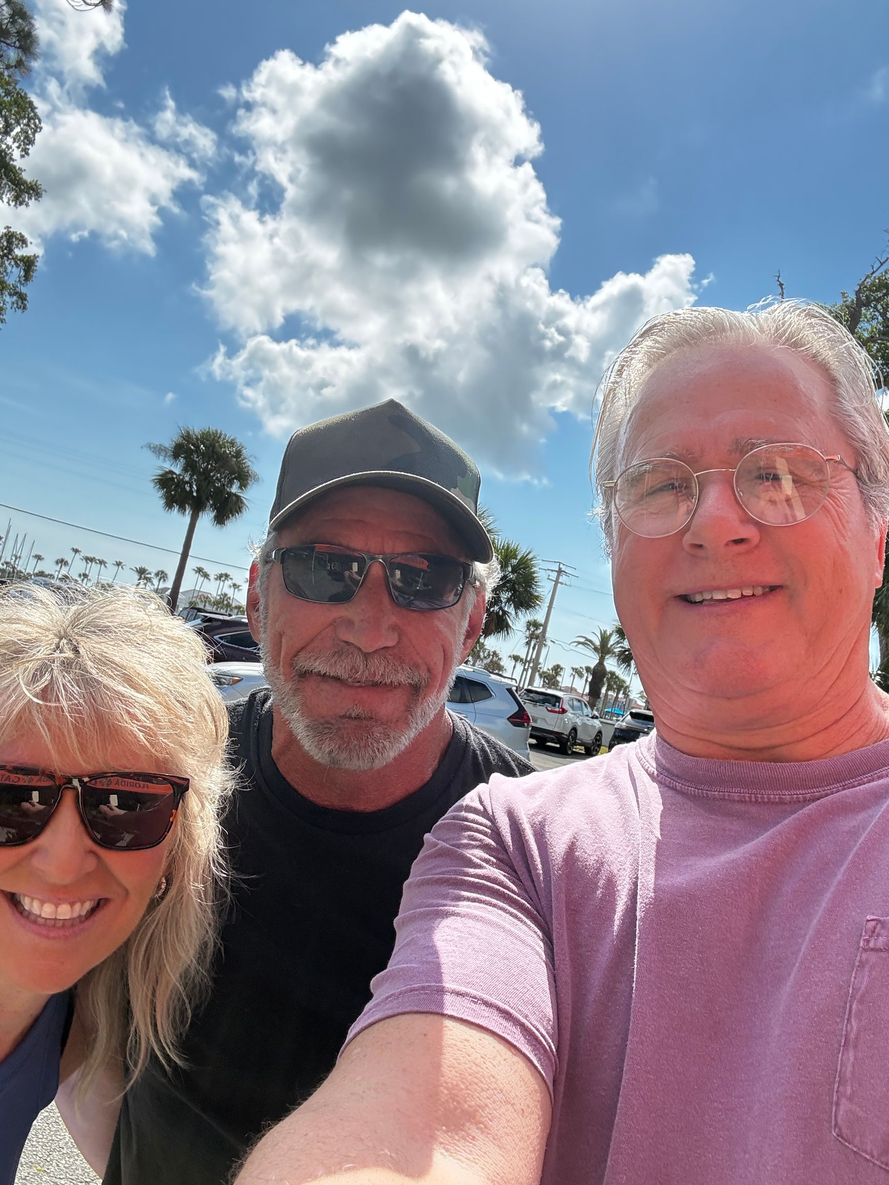 Three smiling people taking a sunny outdoor selfie under a blue sky with clouds.