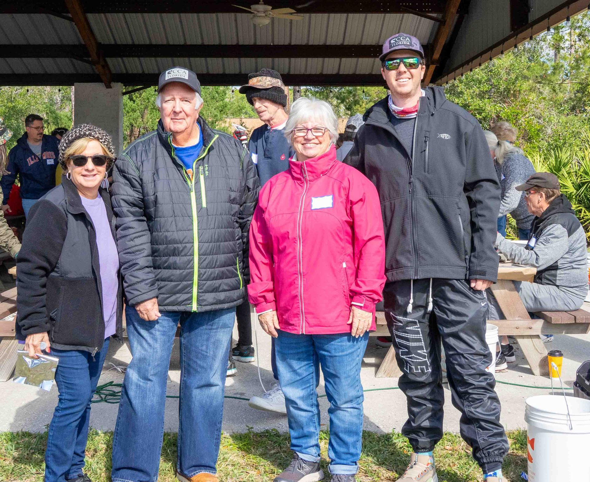 Four people in jackets stand outdoors under a pavilion, smiling at a casual gathering.