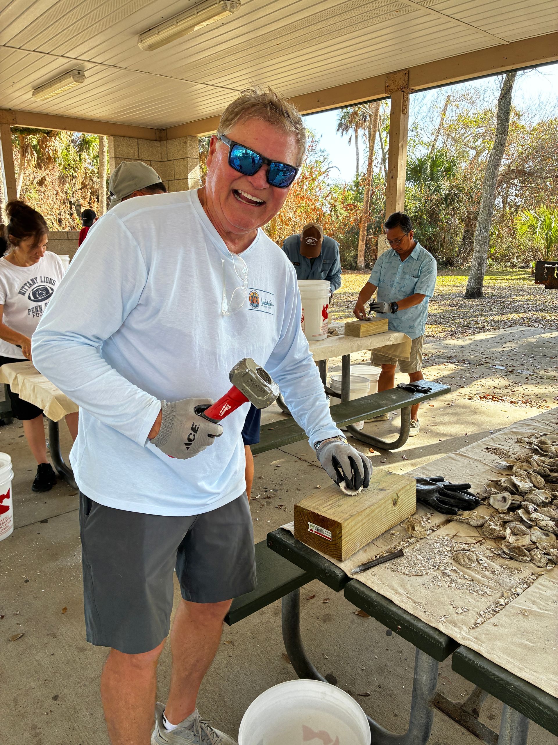 Smiling man in blue sunglasses holding a hammer at an outdoor workbench under a shelter