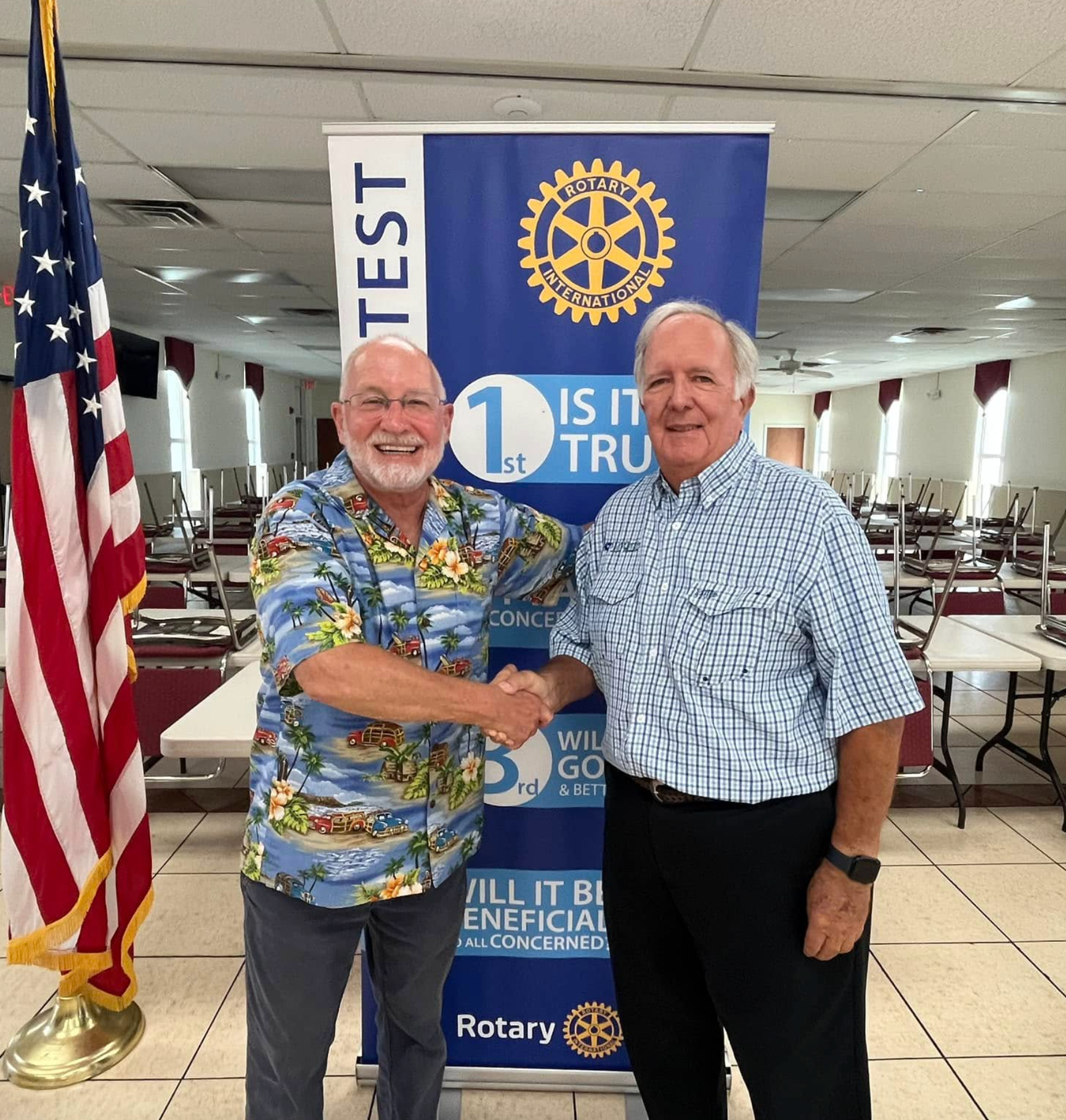Two men shaking hands in front of a Rotary banner at an indoor event