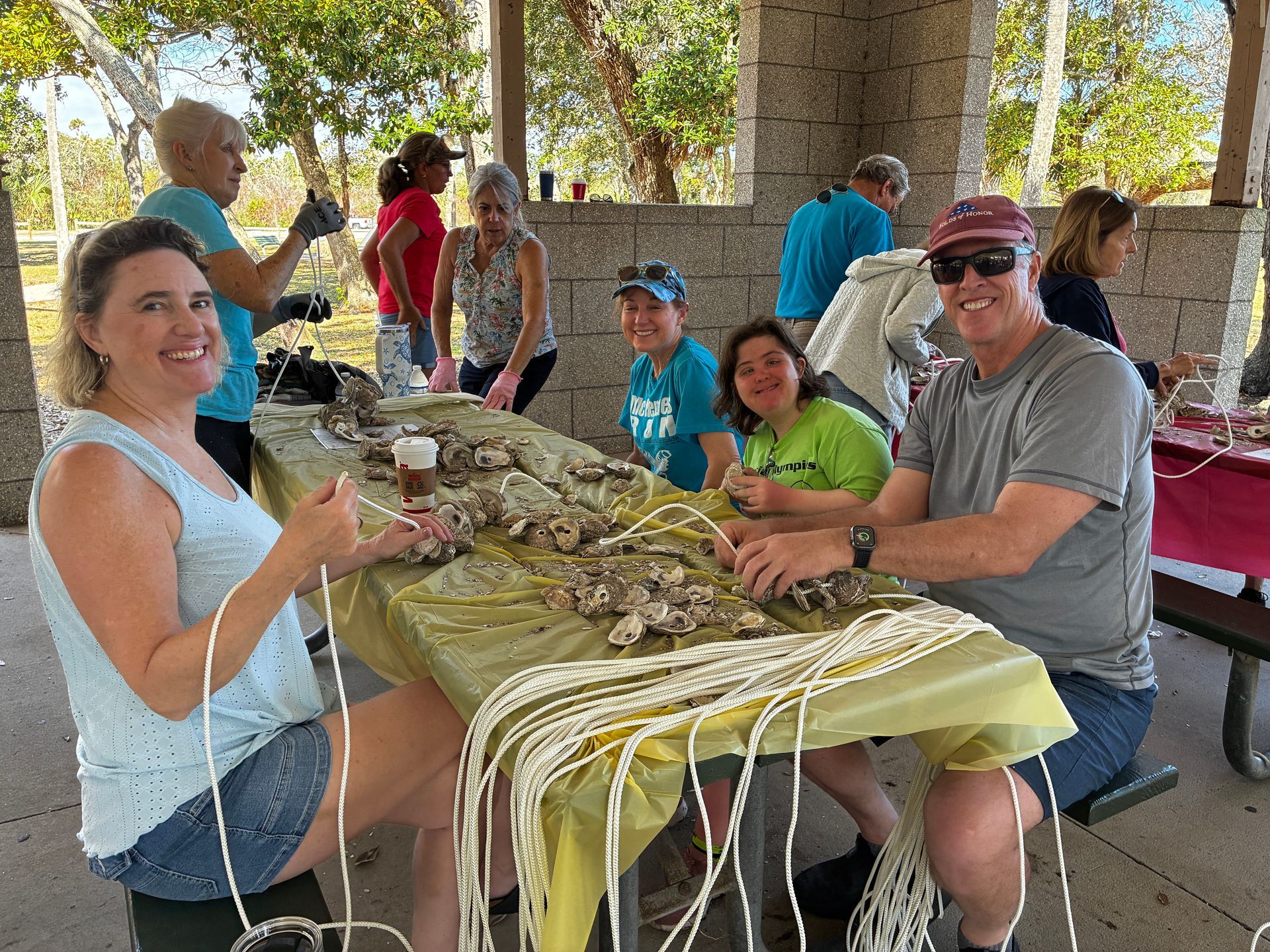 People seated at a picnic table sharing a meal under a pavilion, with others in the background.