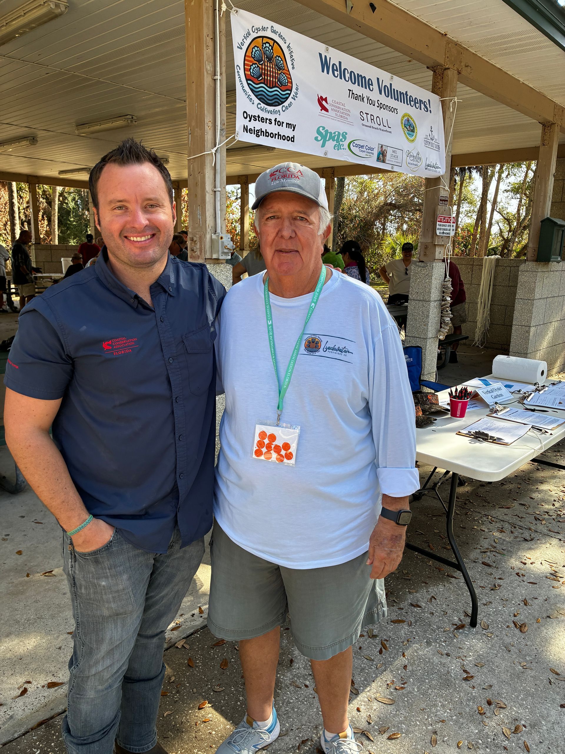 Two men standing under a booth canopy at an outdoor event, smiling beside a banner and table.