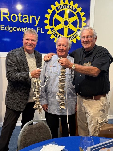 Three men pose holding a long garland at a Rotary Edgewater, FL event banner