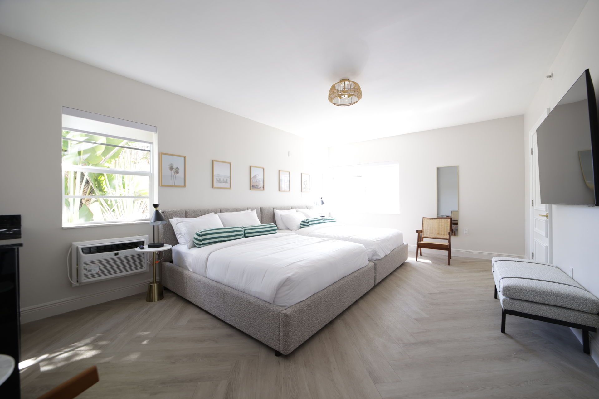 Bright bedroom with two beds, window, artwork, TV, and light wood flooring.