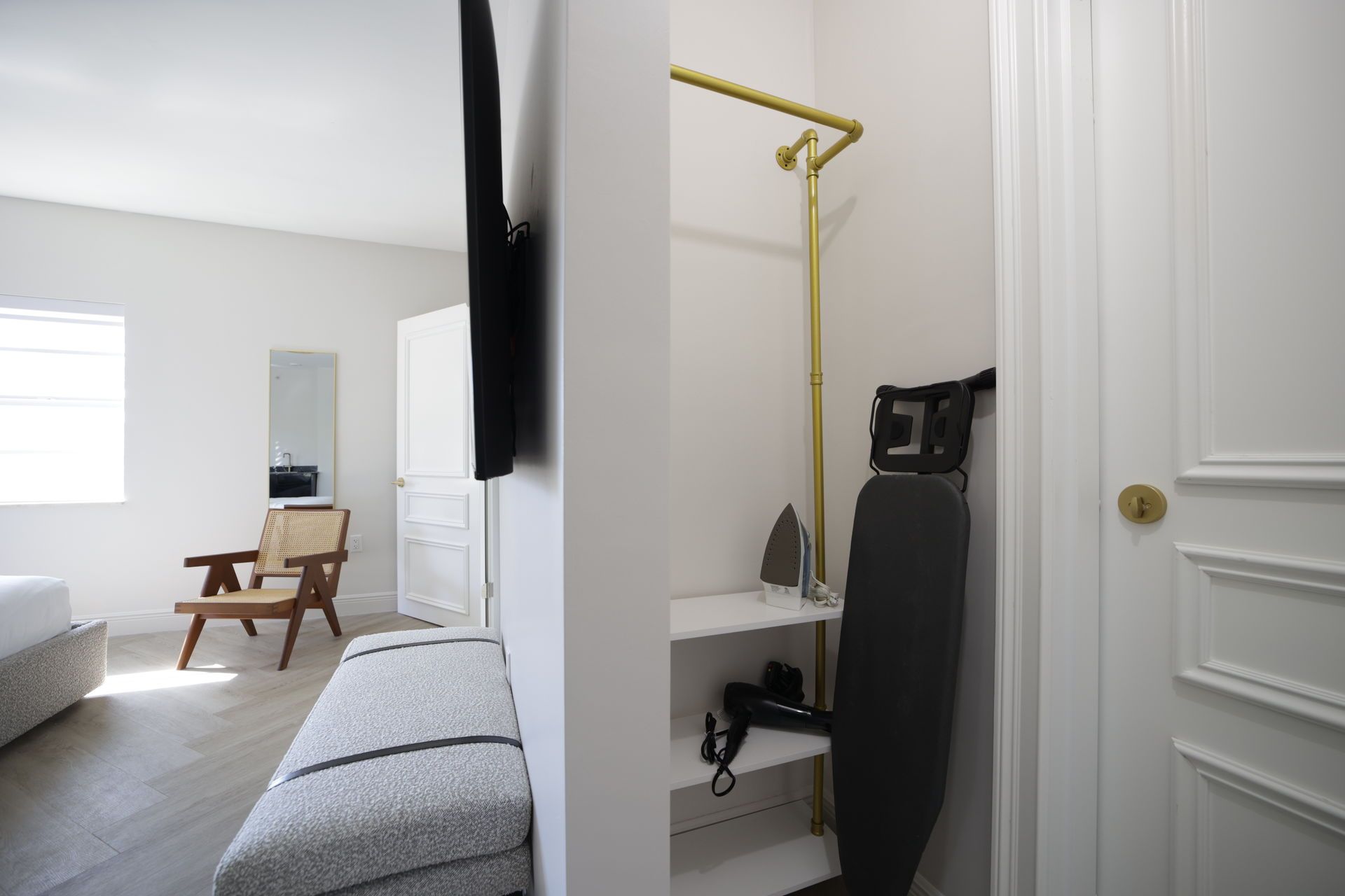Bedroom corner with built-in closet, gold clothing rod, ironing board, neutral colors, modern design.