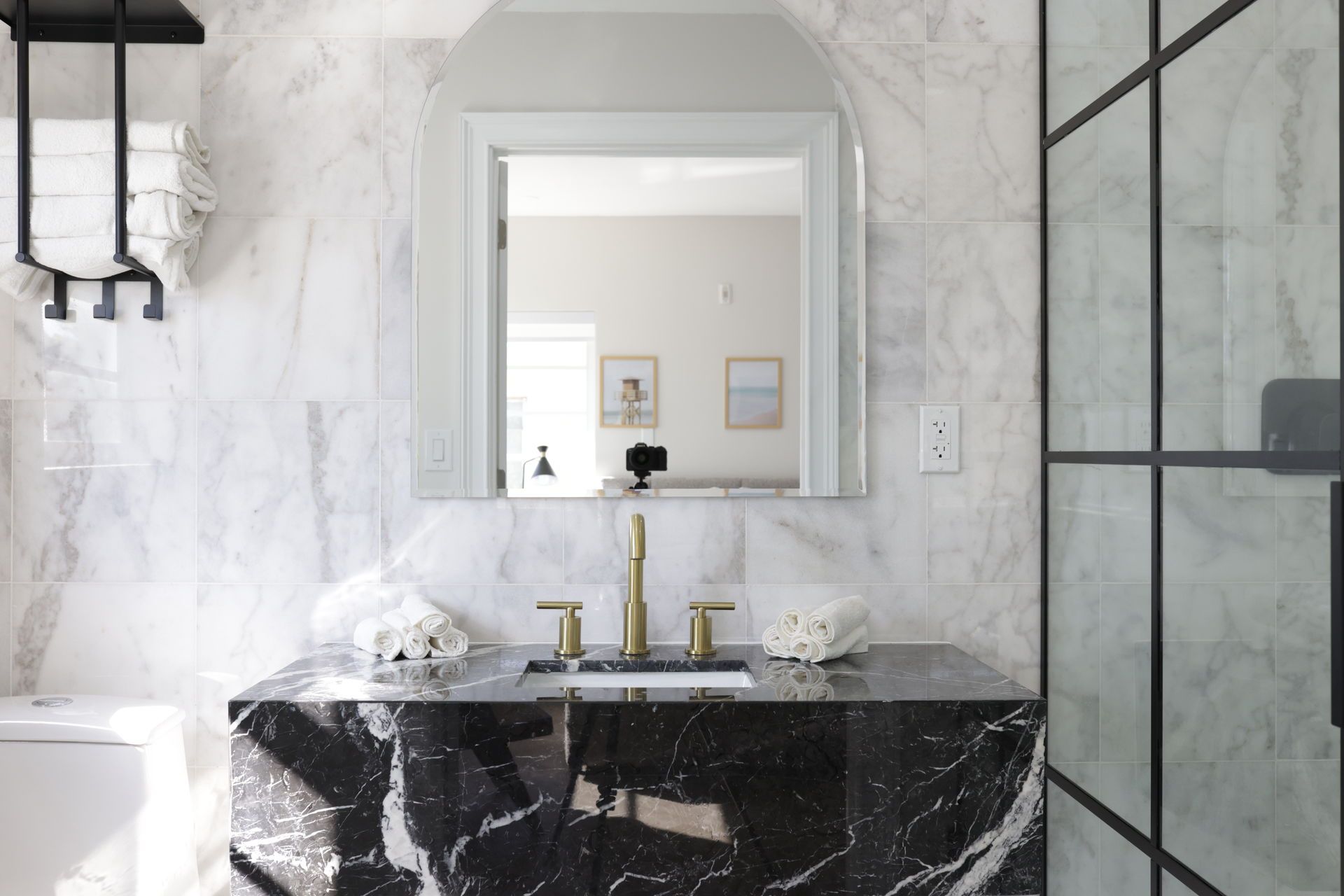 Bathroom with marble walls and a black marble vanity, gold fixtures, arched mirror, and black-framed shower.
