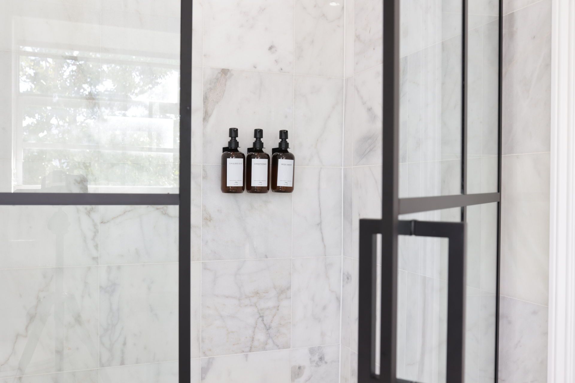 Three dark bottles with white labels mounted on a marble wall inside a shower.