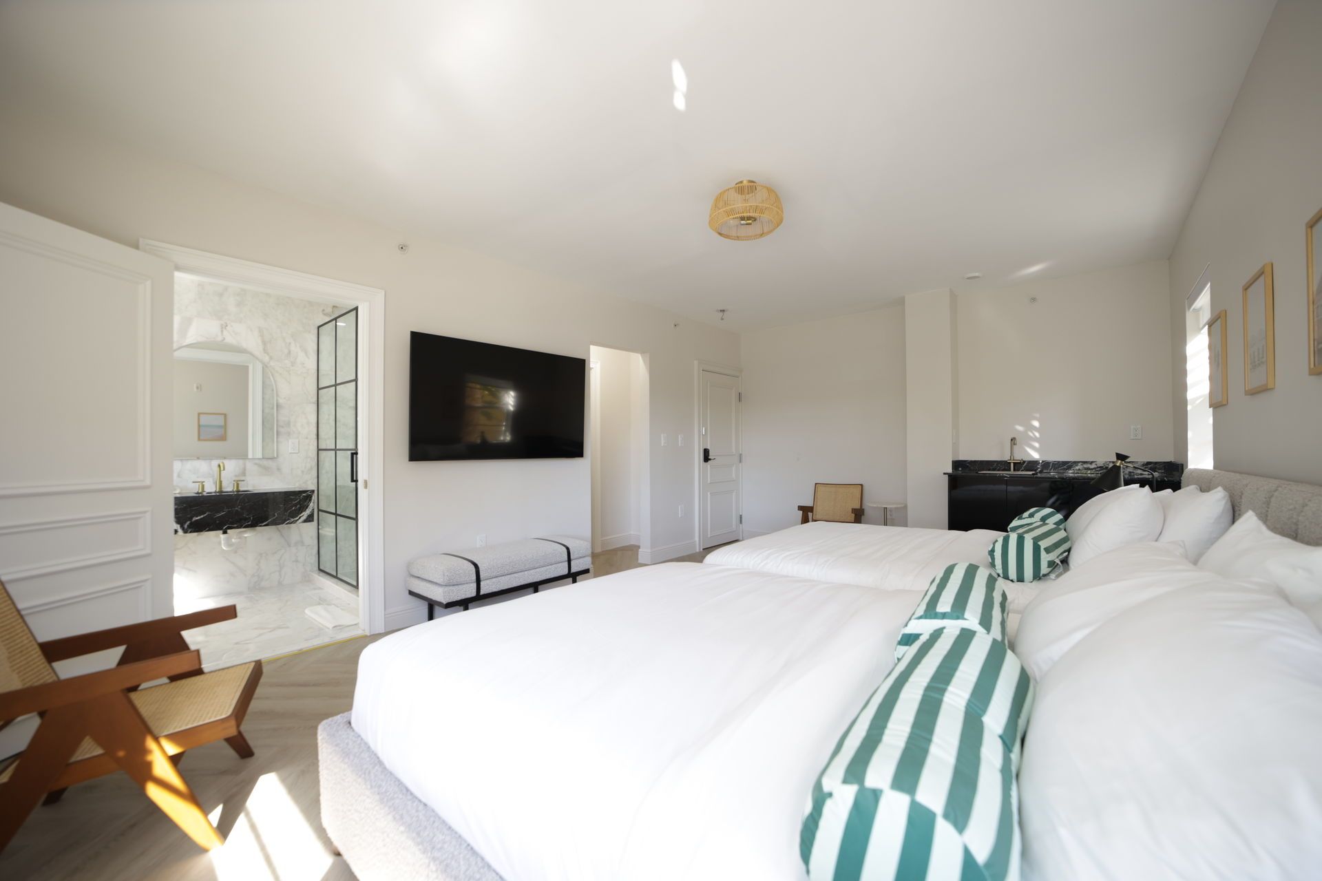 Bright bedroom with two beds, open bathroom, TV, and sunlight.