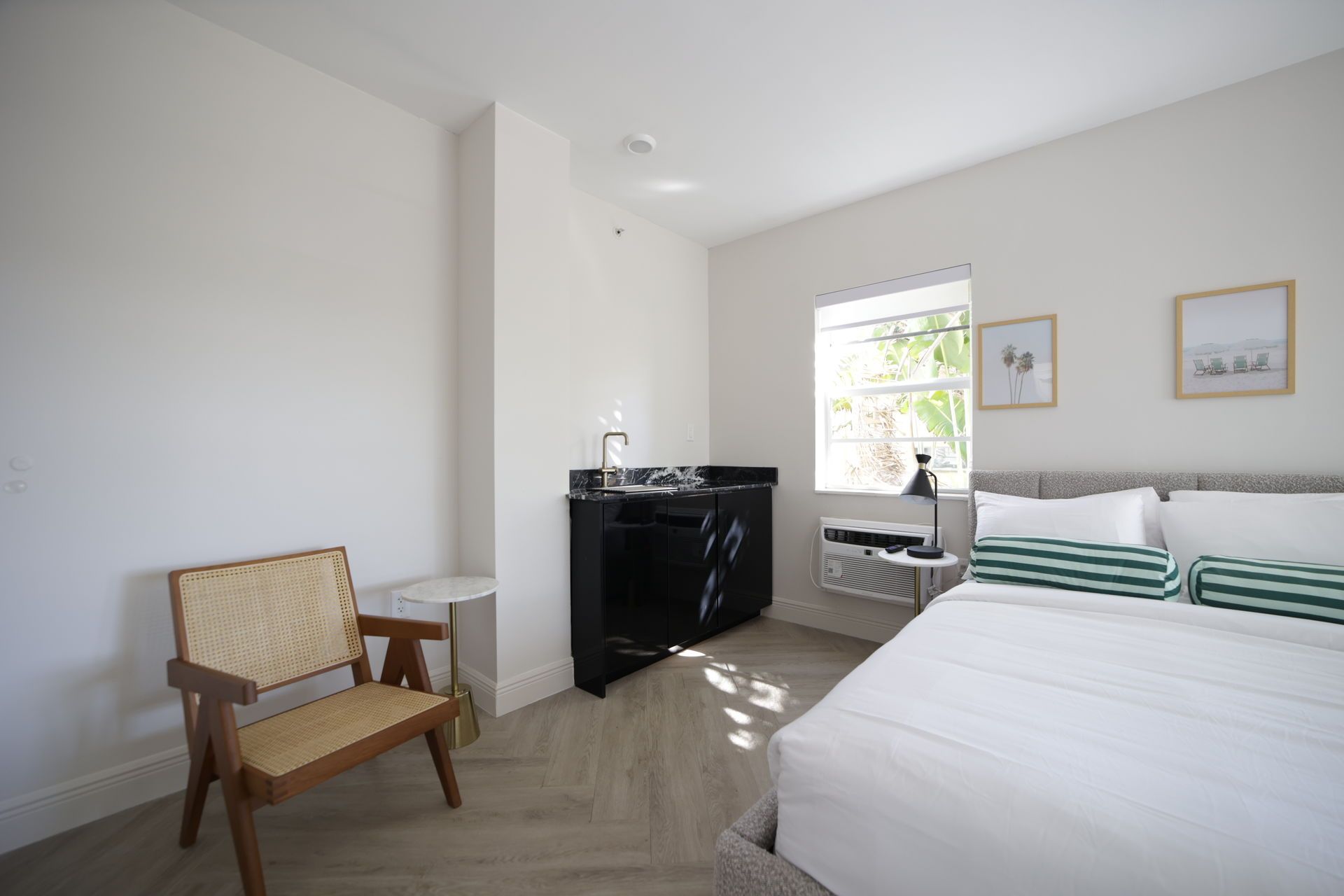 Bright bedroom with a bed, kitchenette, chair, and window. White walls, light wood floor.