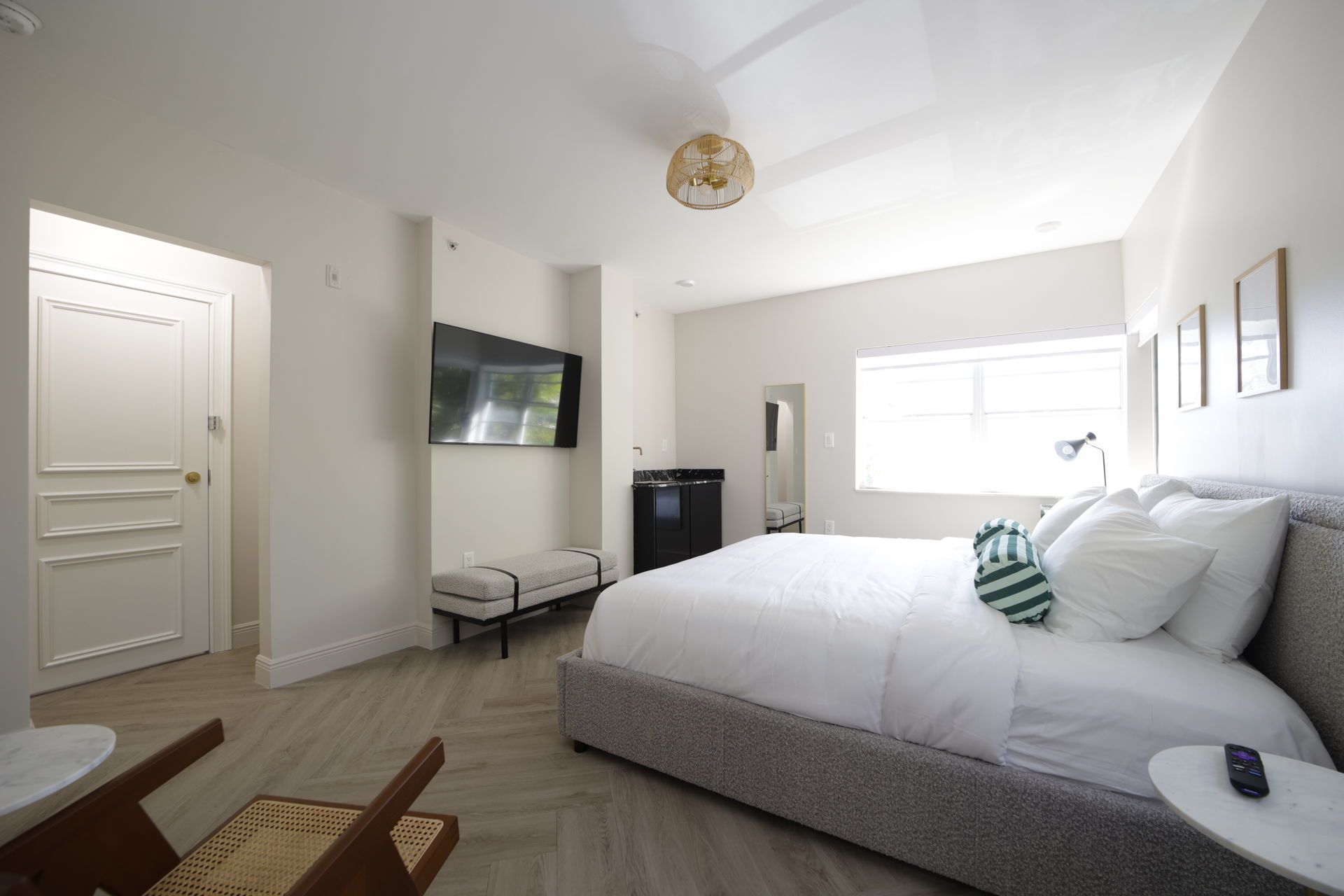 Bright hotel room with king-size bed, TV, mini-fridge, and window. White walls, wooden floor, minimalist decor.