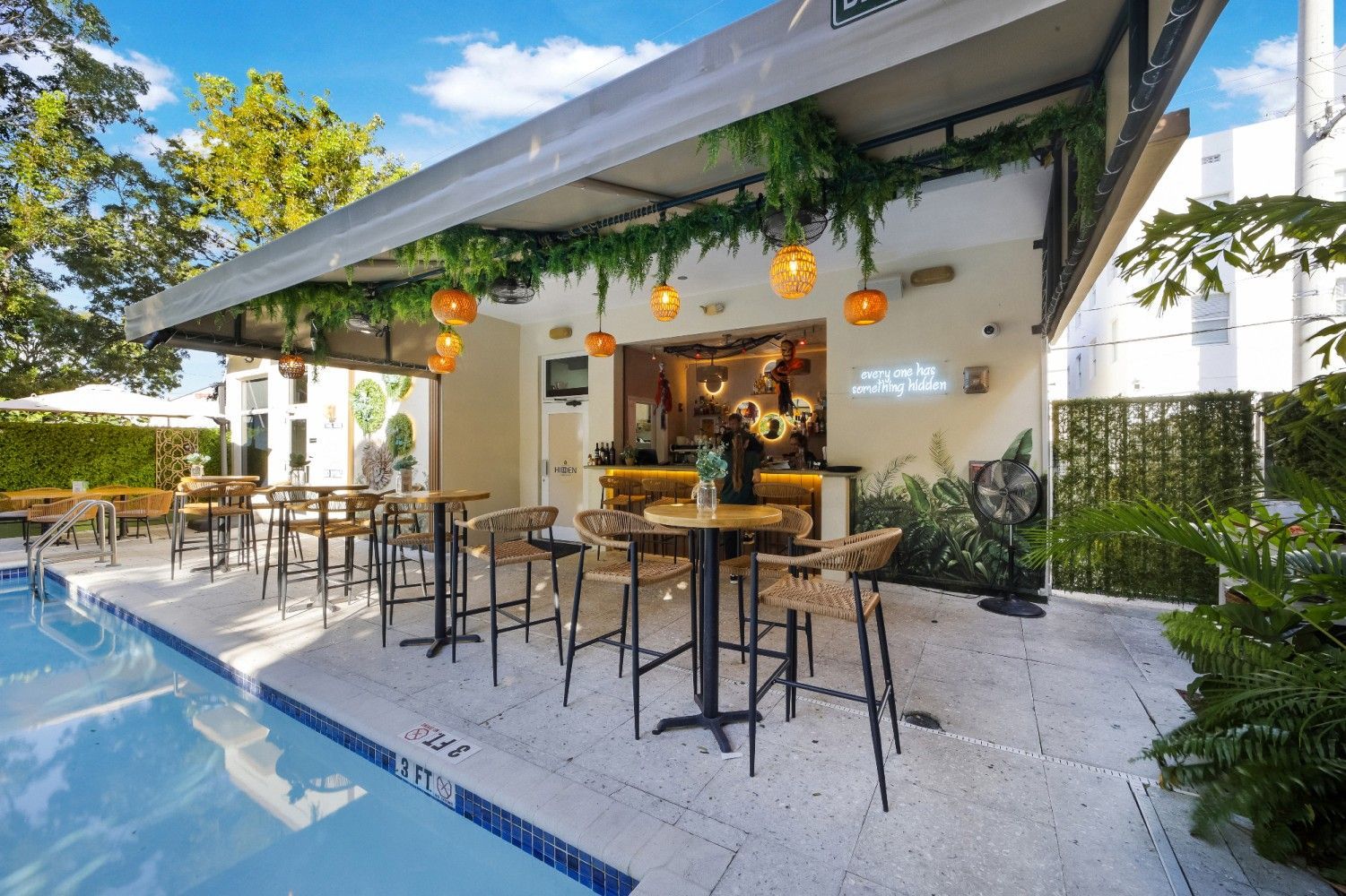 Poolside bar with high-top tables and stools. Covered bar with hanging lights and greenery.