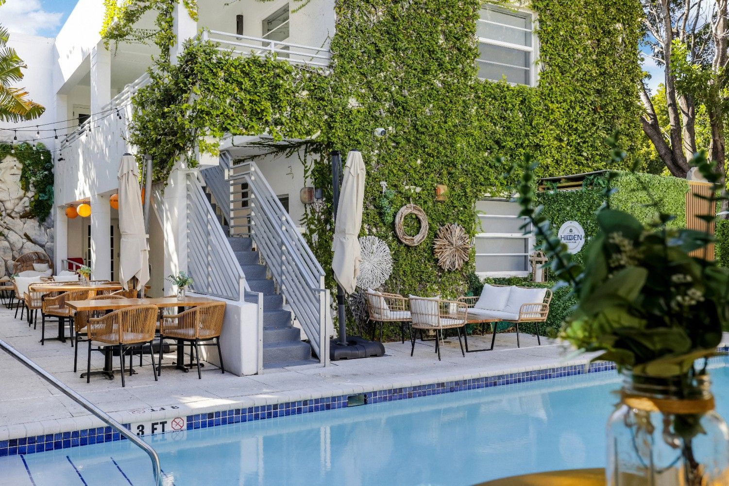 Outdoor hotel restaurant with pool, white building with green vines, wooden tables and chairs.
