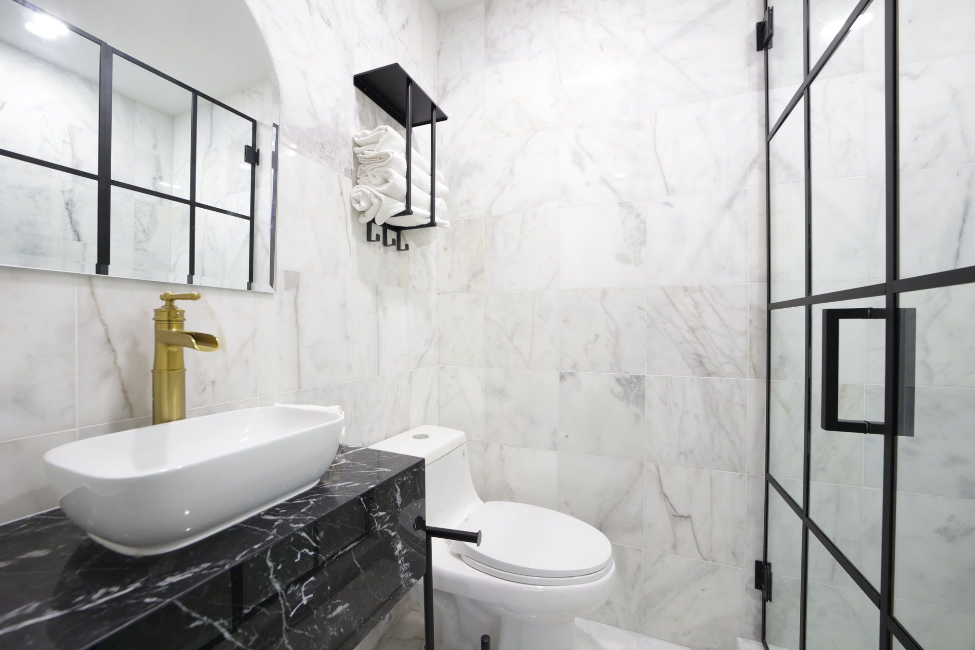 Modern bathroom with marble walls, a gold faucet, black vanity, and a glass shower door.