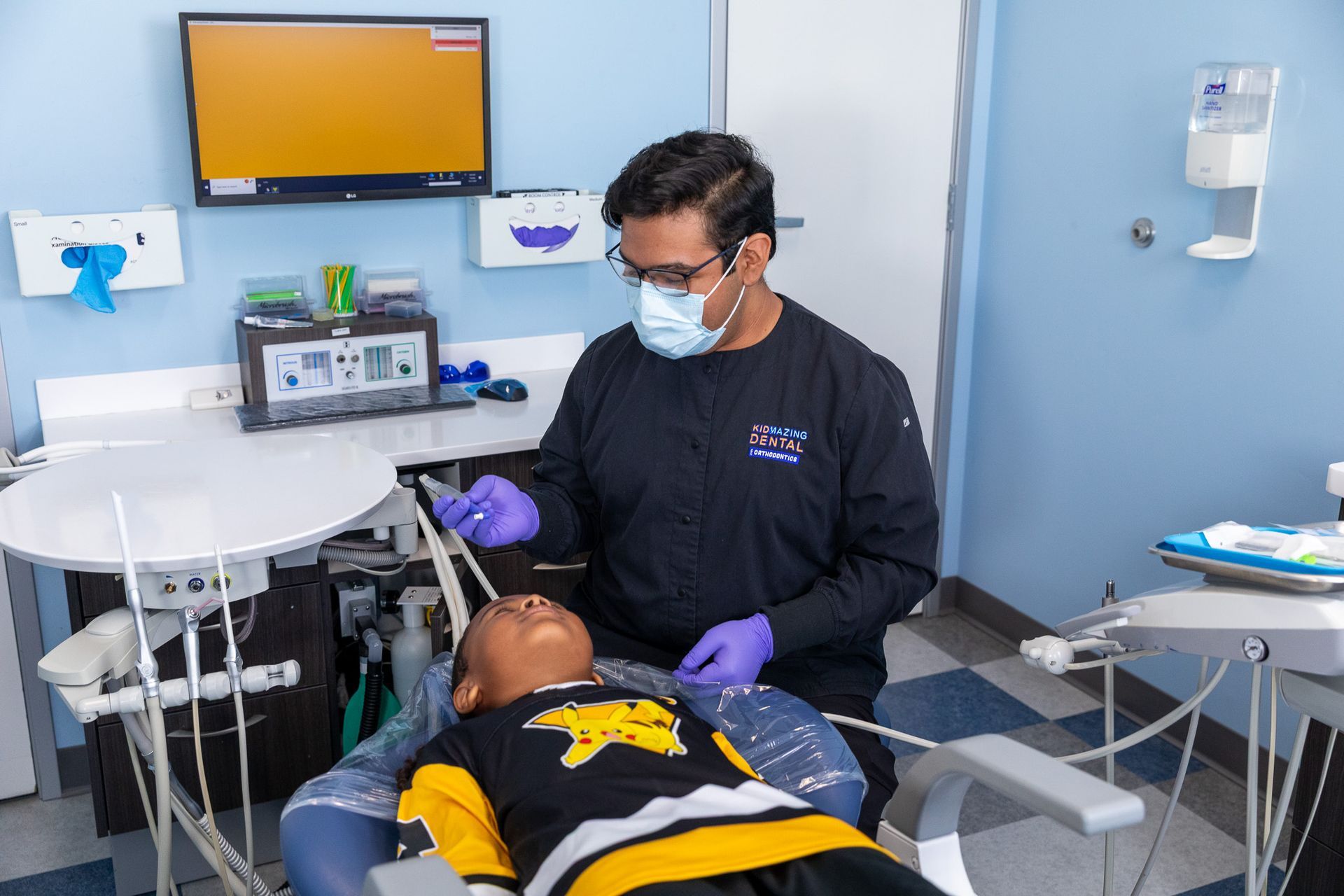 Dentist treating a patient in a clinic, with medical equipment and a monitor nearby.