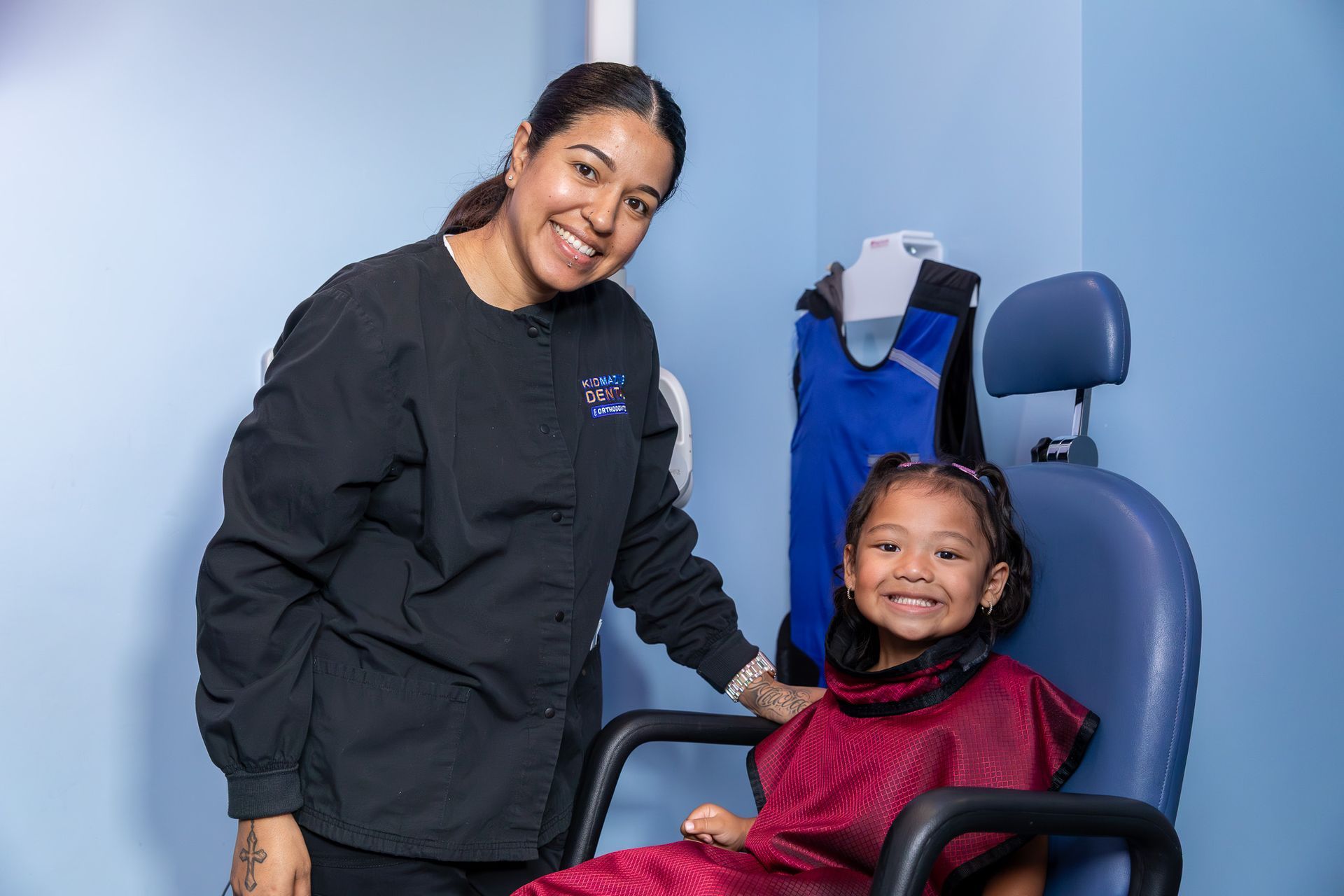 Dental assistant and child smiling in a dental office; child in chair, wearing protective gear.