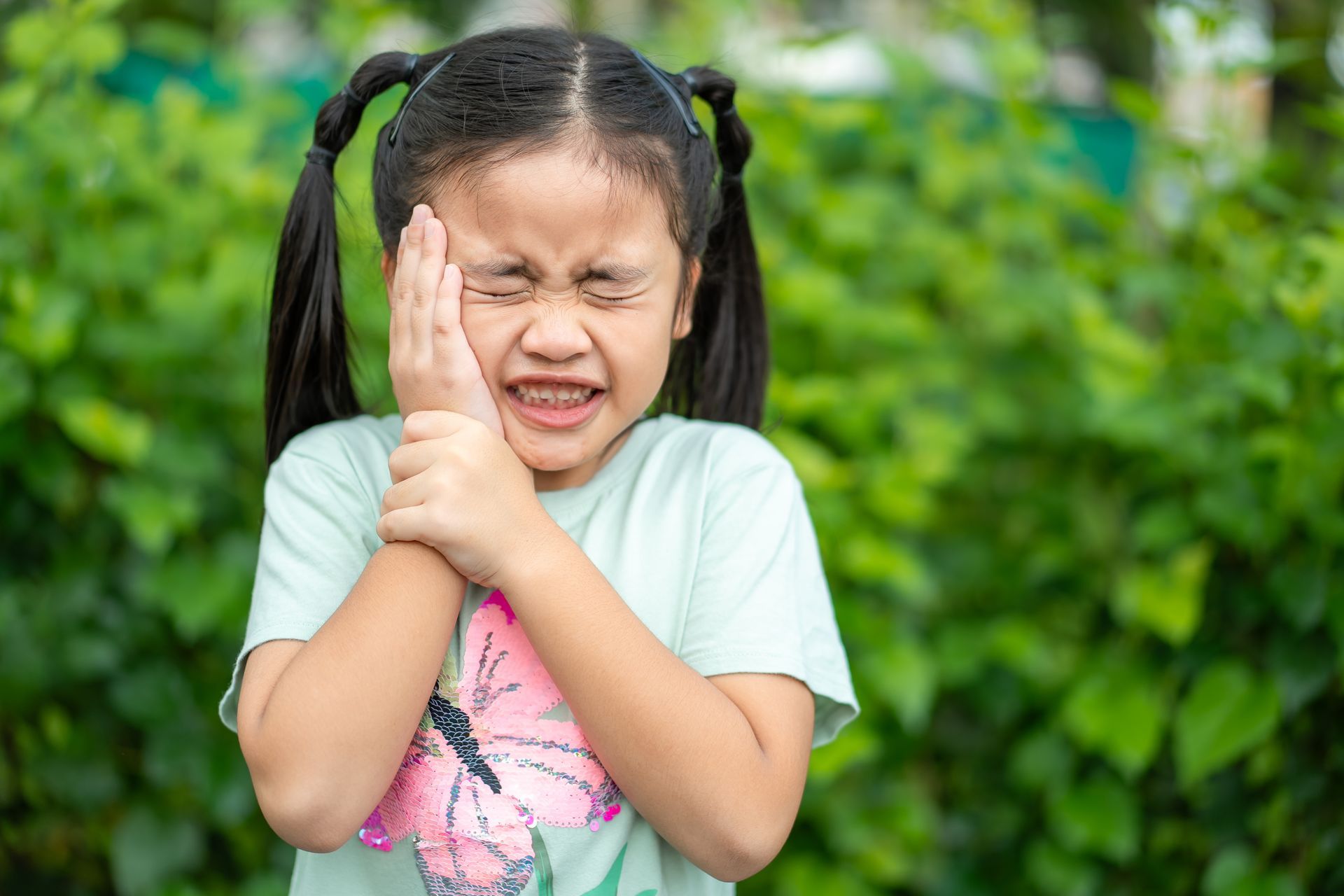 Child grimacing and clutching their arm against a green leafy background