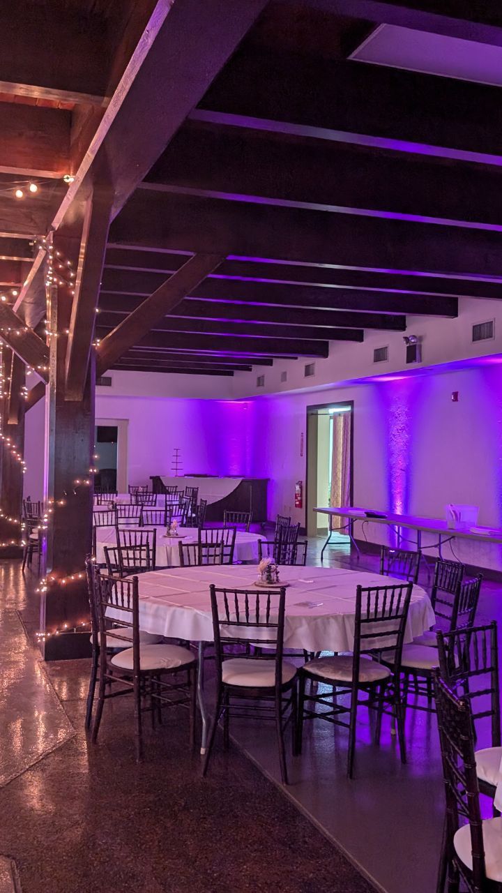 Purple-lit banquet hall with round tables, black chairs, and exposed ceiling beams