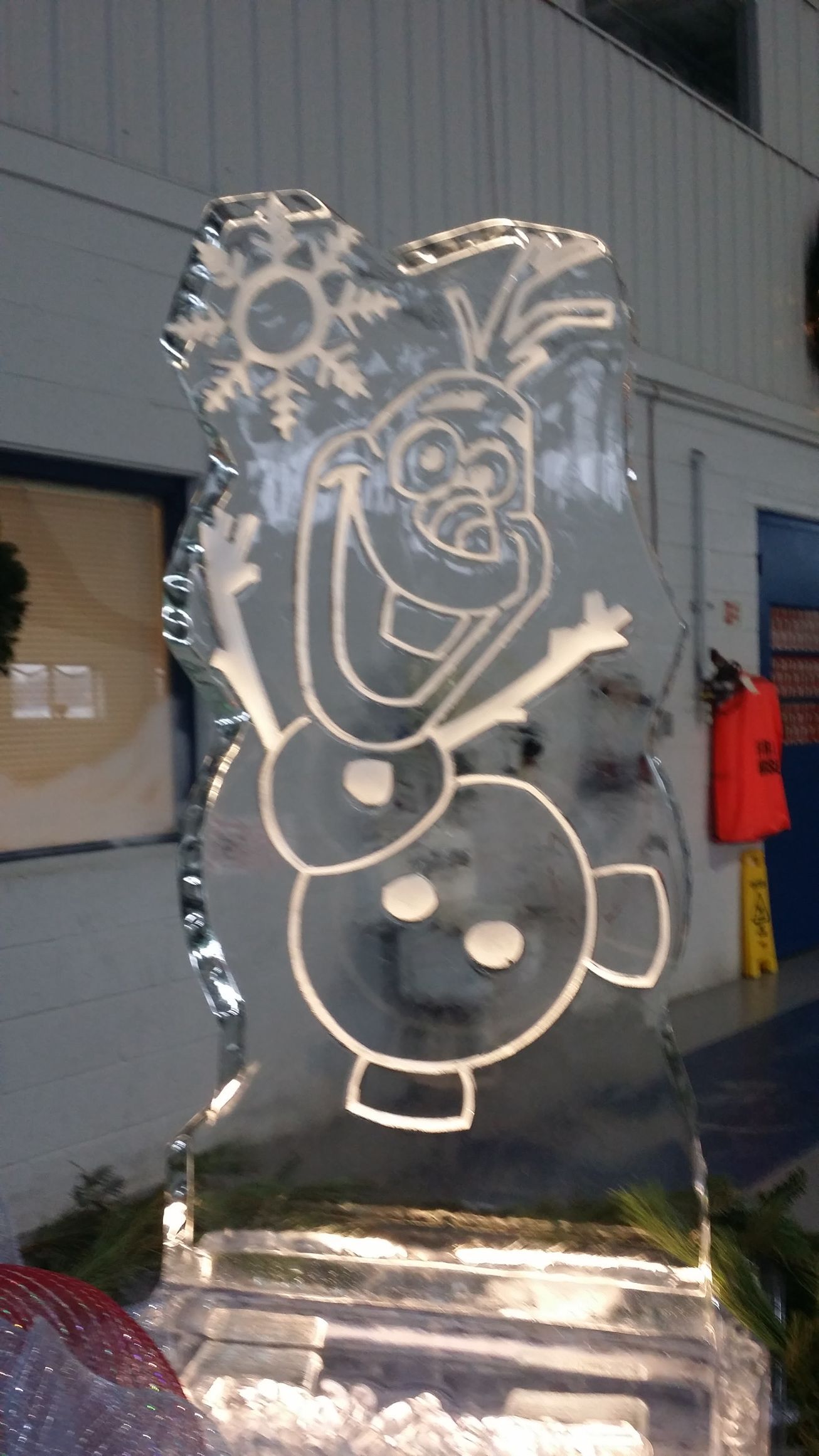 Transparent ice sculpture of Olaf from Frozen, lit indoors against a building wall.