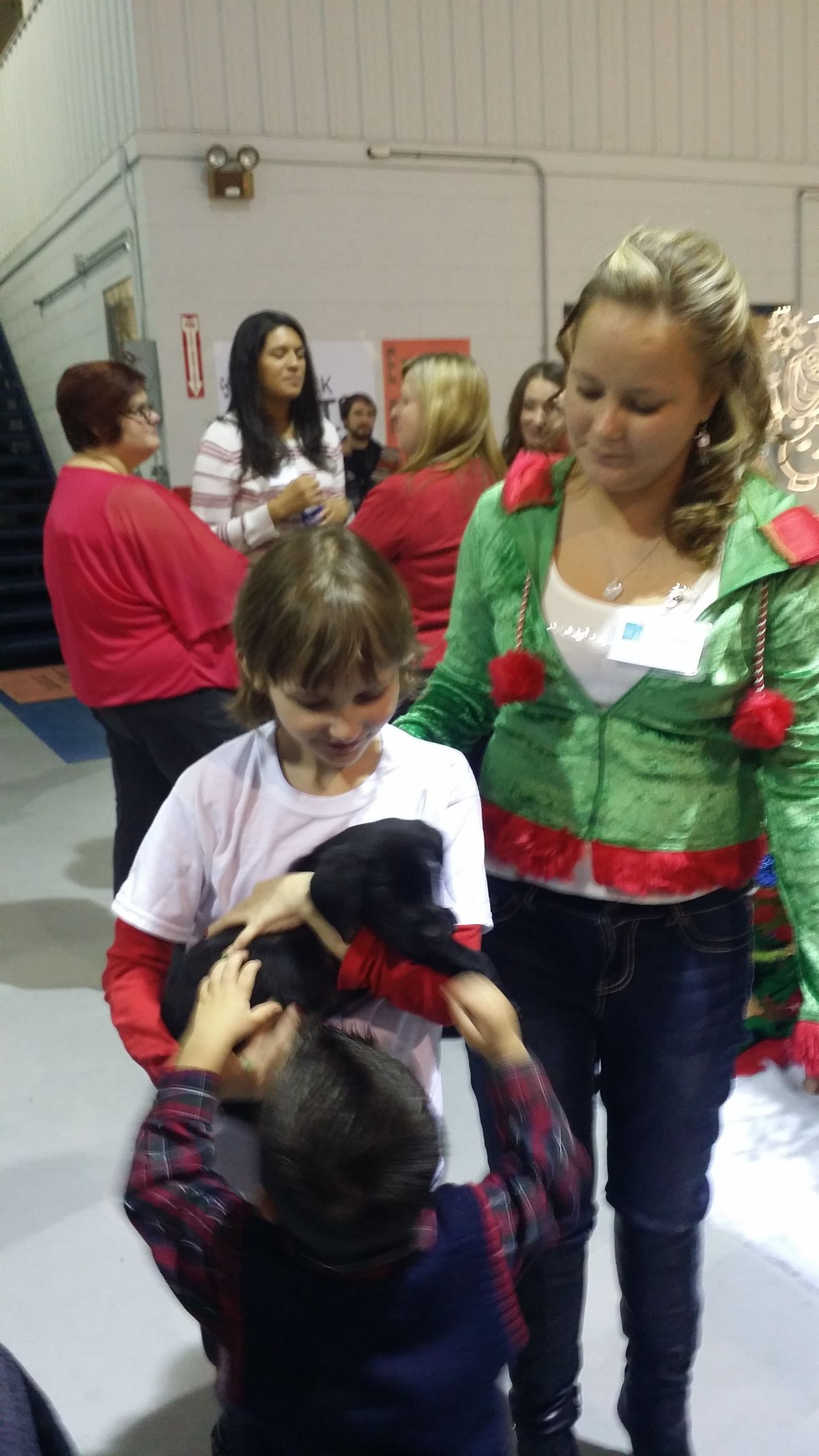Two children in a gym, one handing a plush toy to the other while a woman stands nearby.