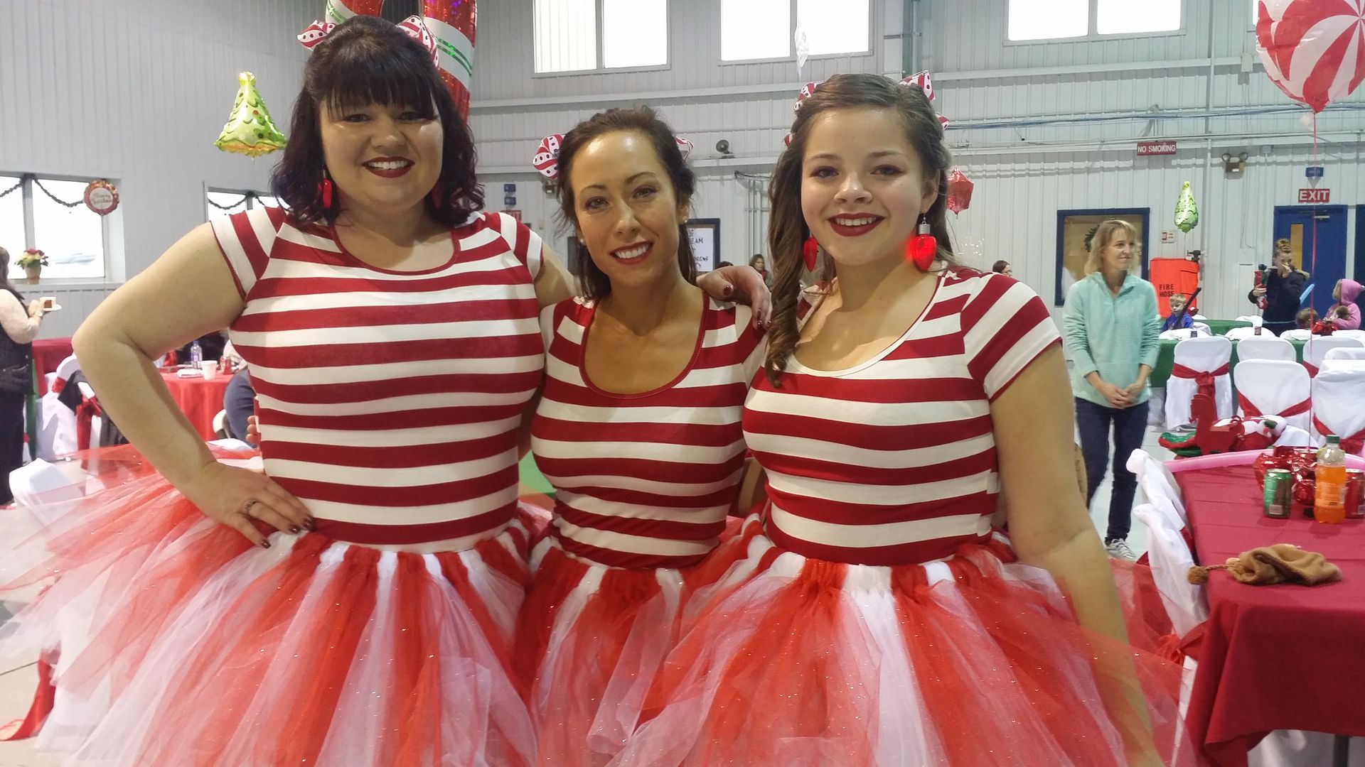 Three performers in red-and-white striped tops and pink tutus smiling at an indoor event